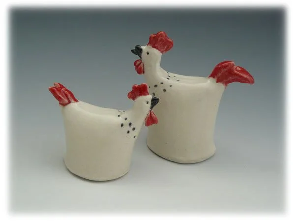 Ceramic chicken and rooster figurines, white with red combs and wattles, and black beaks, set against a plain background.