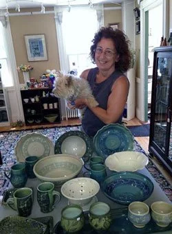 Woman holding a small dog standing behind a table filled with various green, blue, and white ceramic bowls and cups in a bright room.