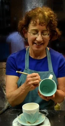 A woman with curly hair and glasses, wearing a blue shirt and apron, is painting a ceramic cup with a brush in a studio.