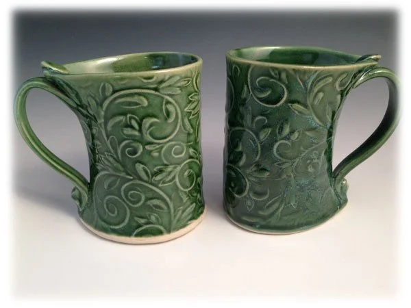 Two green ceramic mugs with spiral and floral patterns on a light background.