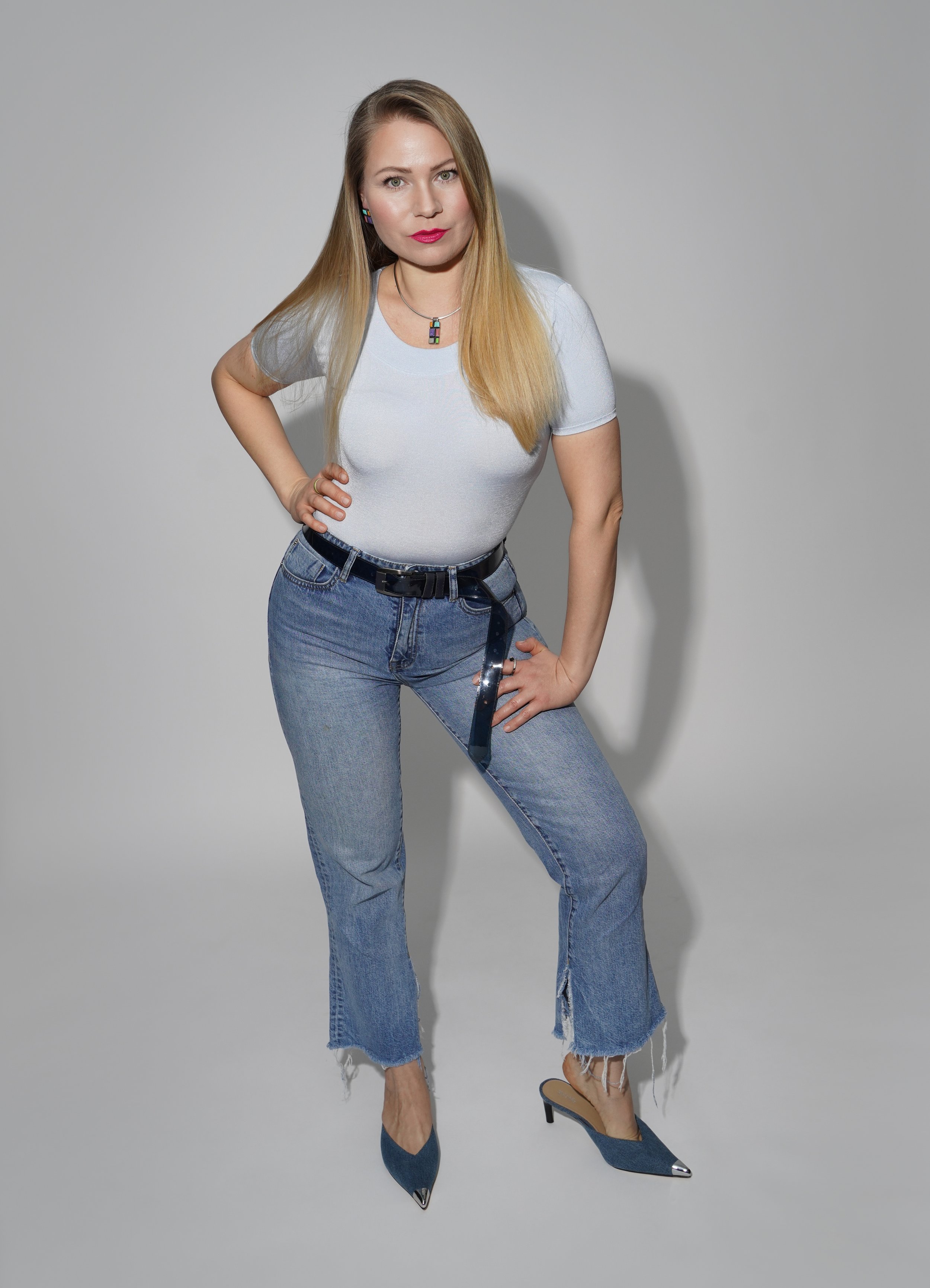 A woman with long blonde hair, wearing a light gray t-shirt, blue jeans, and blue high-heeled shoes, standing against a plain gray background with one hand on her hip and the other resting on her thigh.