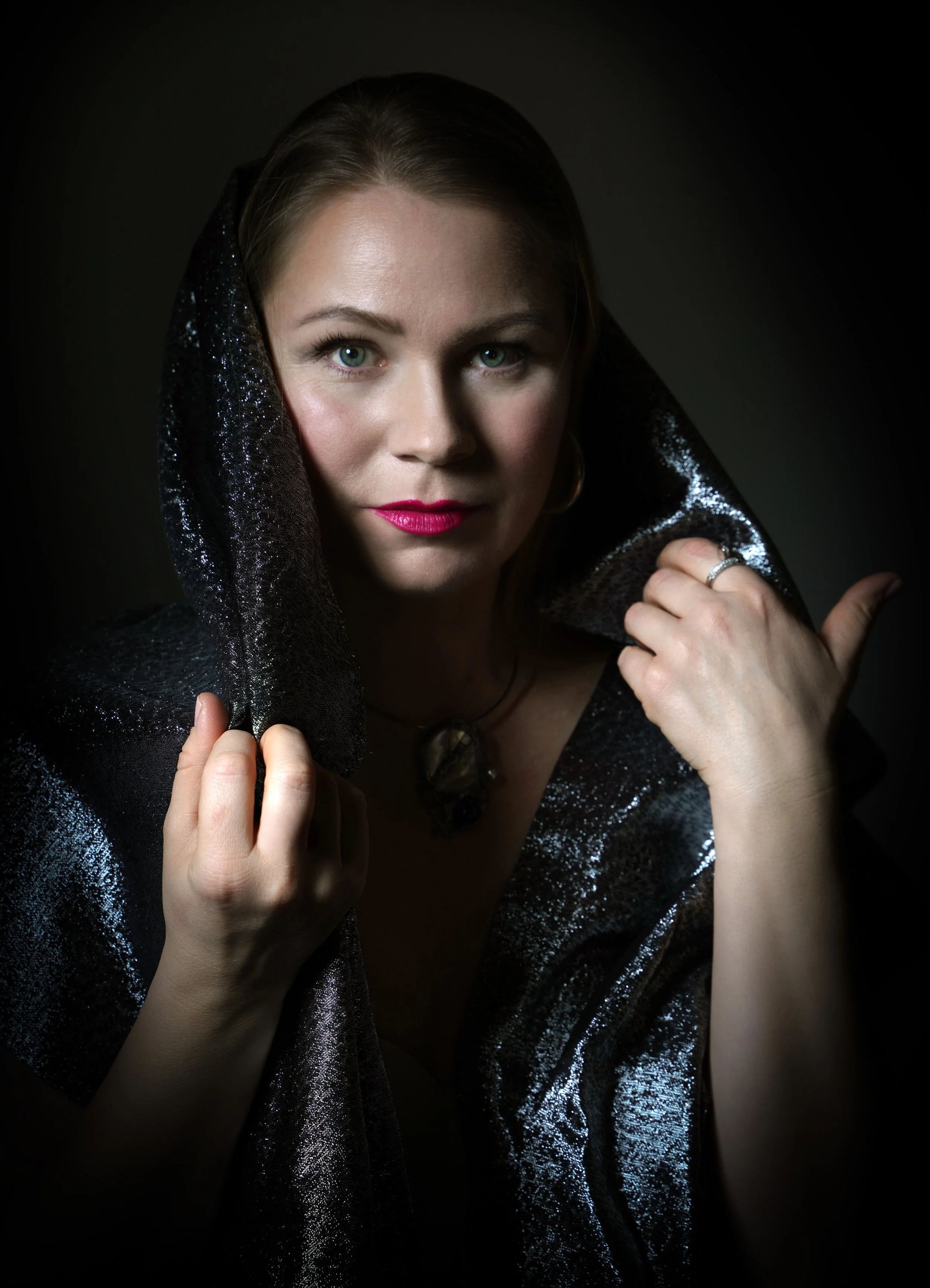 Close-up of a woman with blue eyes and pink lipstick, wearing a shimmering black hooded shawl, against a dark background.