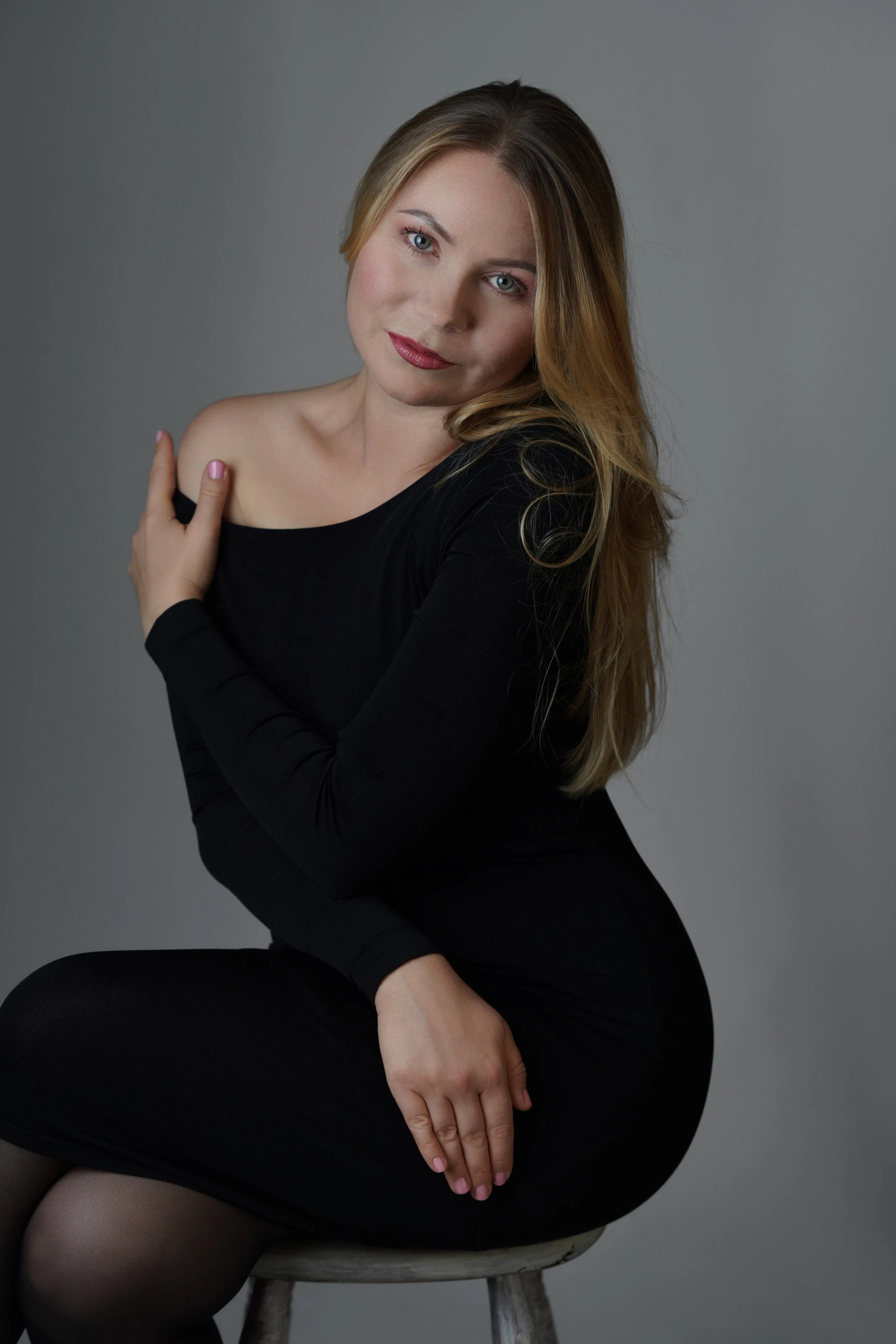 A woman with long blonde hair, wearing a black top and tights, sitting on a wooden stool against a plain gray background, looking at the camera with a gentle expression.