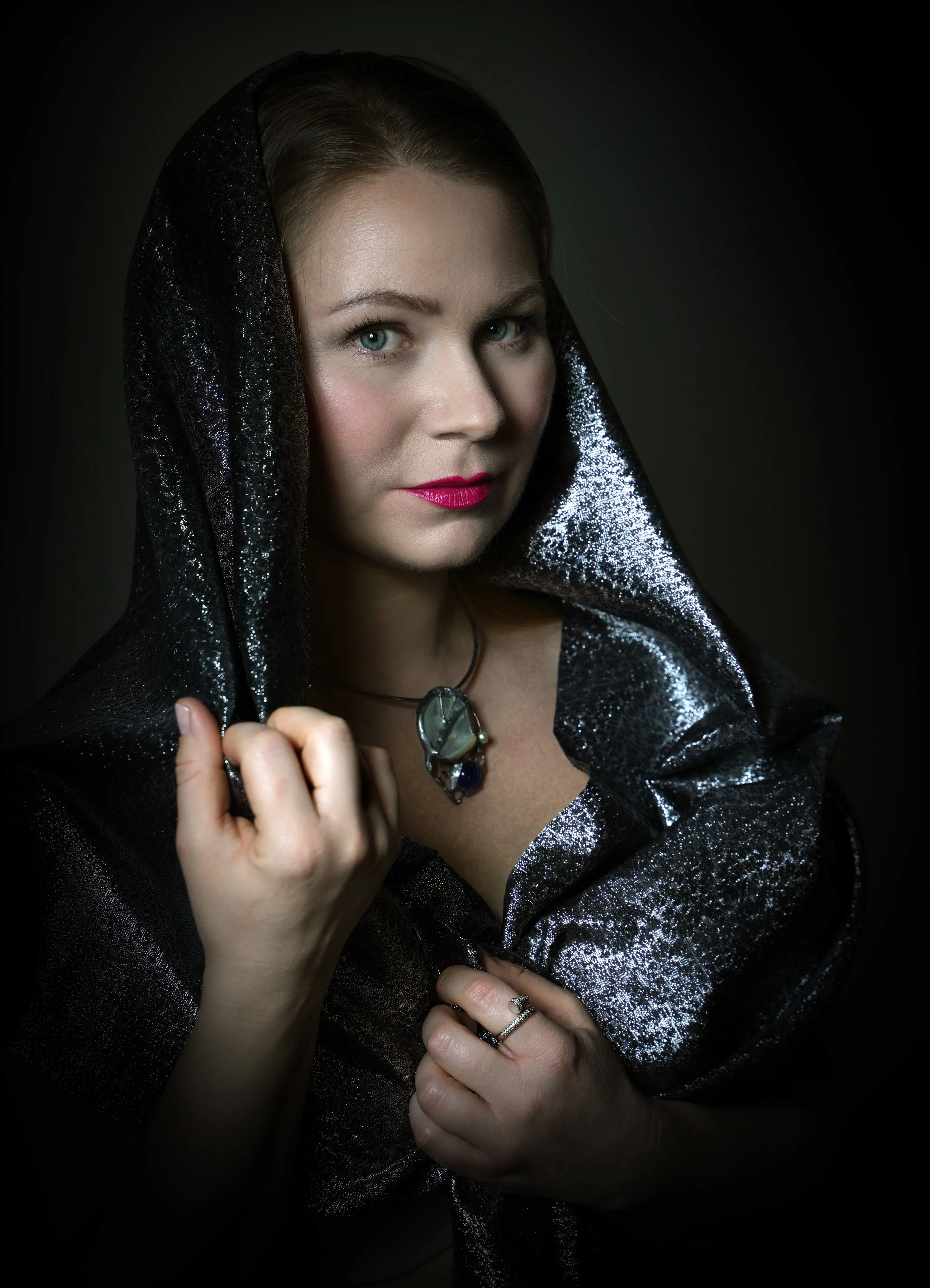 A woman with green eyes and pink lipstick, wearing a black, shiny, textured shawl, holding it with her right hand, and wearing a necklace with a large pendant and rings on her fingers.