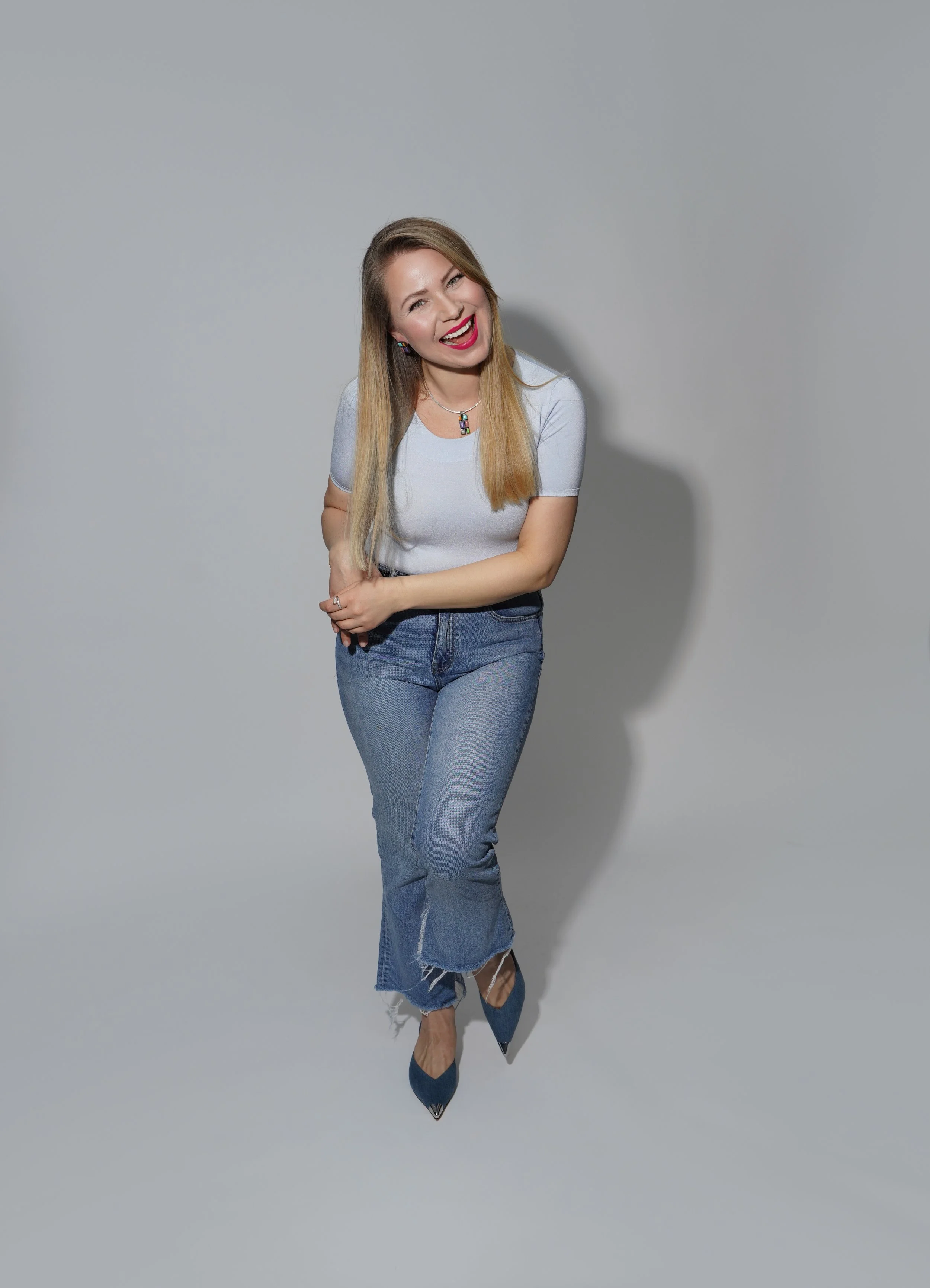 A woman with long, blonde hair wearing a light gray top, blue jeans, and blue heels, standing against a plain gray background, smiling and looking at the camera.