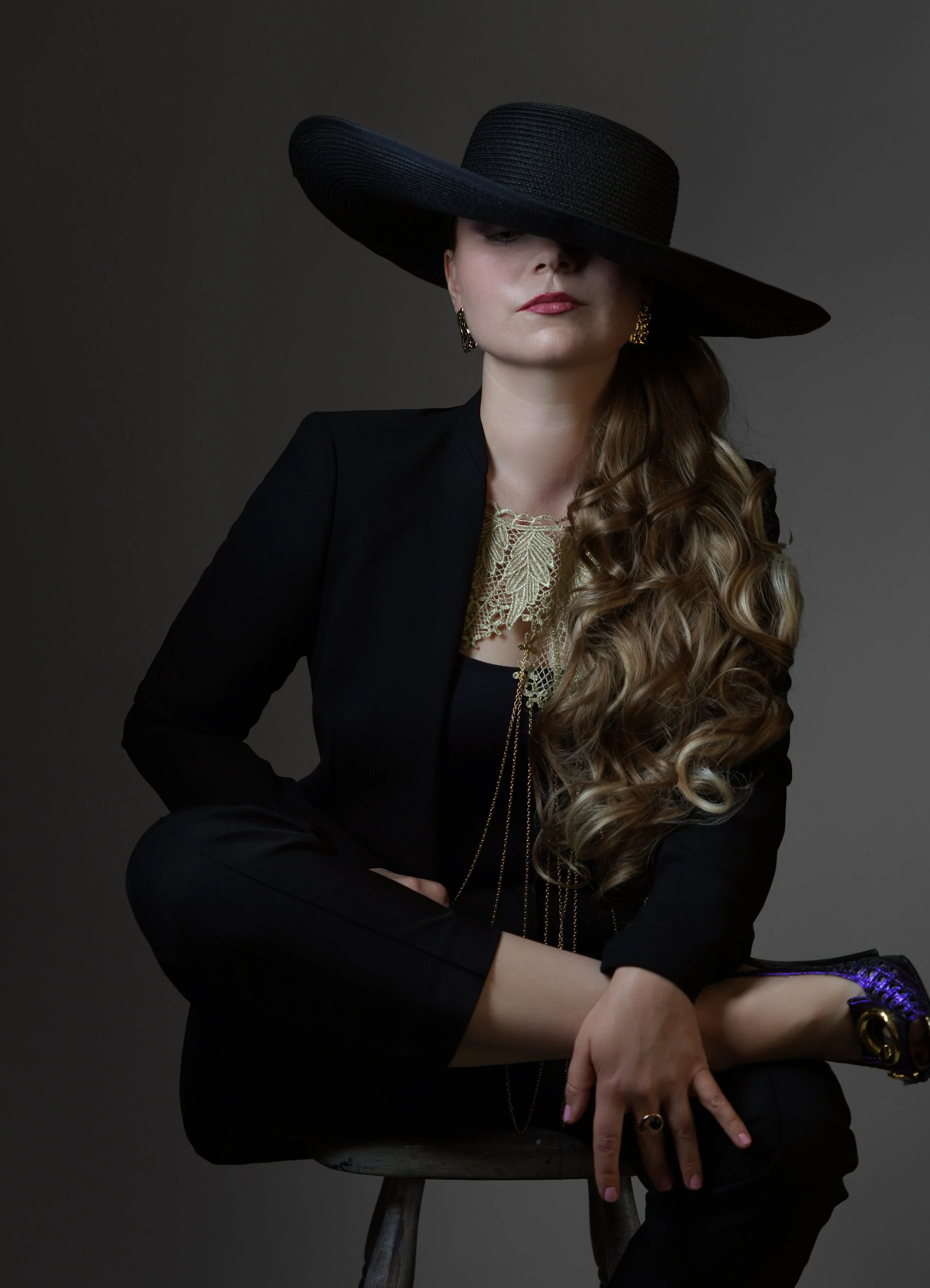 A woman sitting on a stool wearing a large black wide-brim hat covering part of her face, a black blazer, gold jewelry, and black outfit, against a dark gray background.