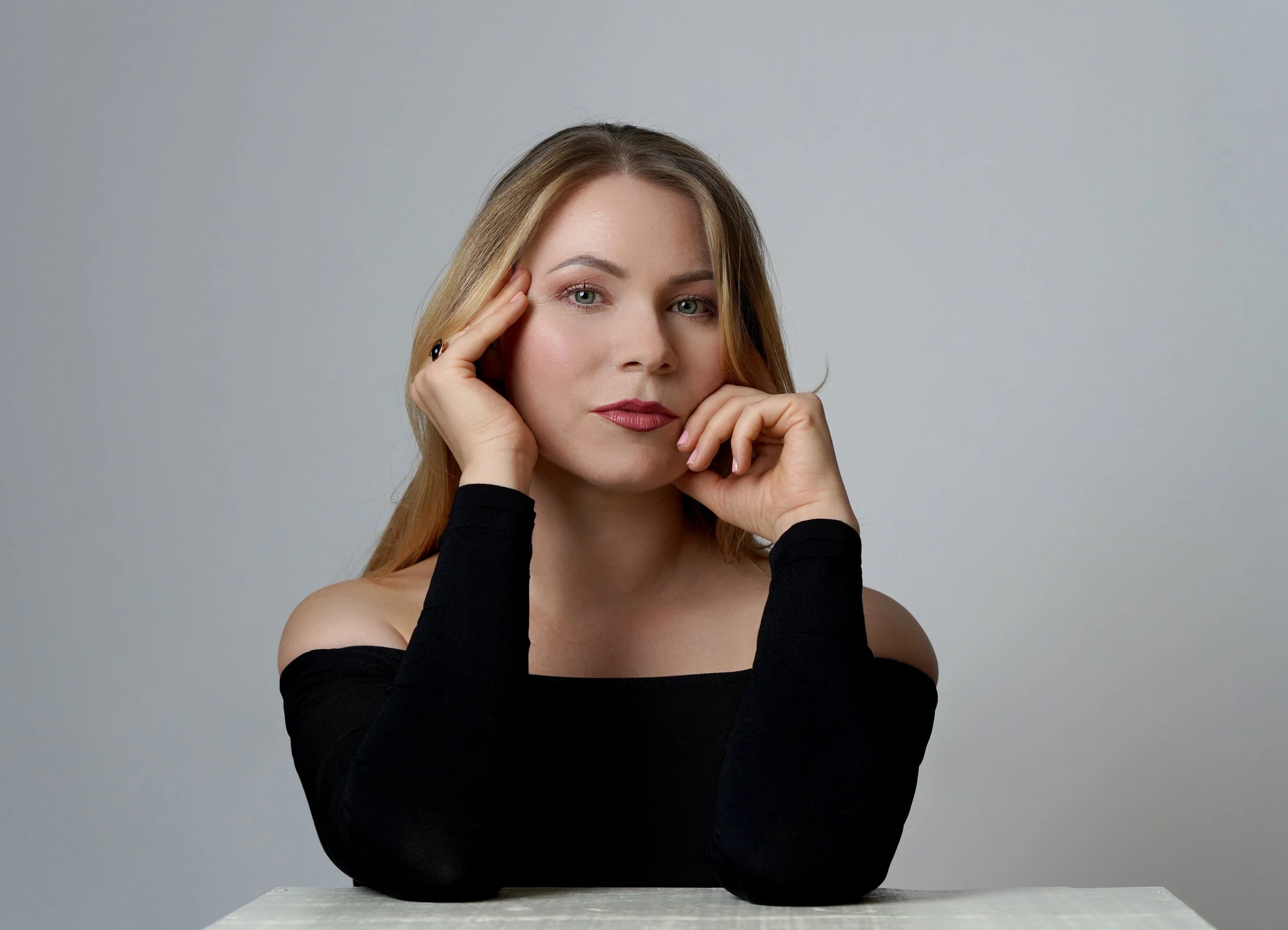A woman with blonde hair and fair skin, resting her face on her left hand and her right hand on her chin, wearing a black off-the-shoulder top, posed against a plain gray background.