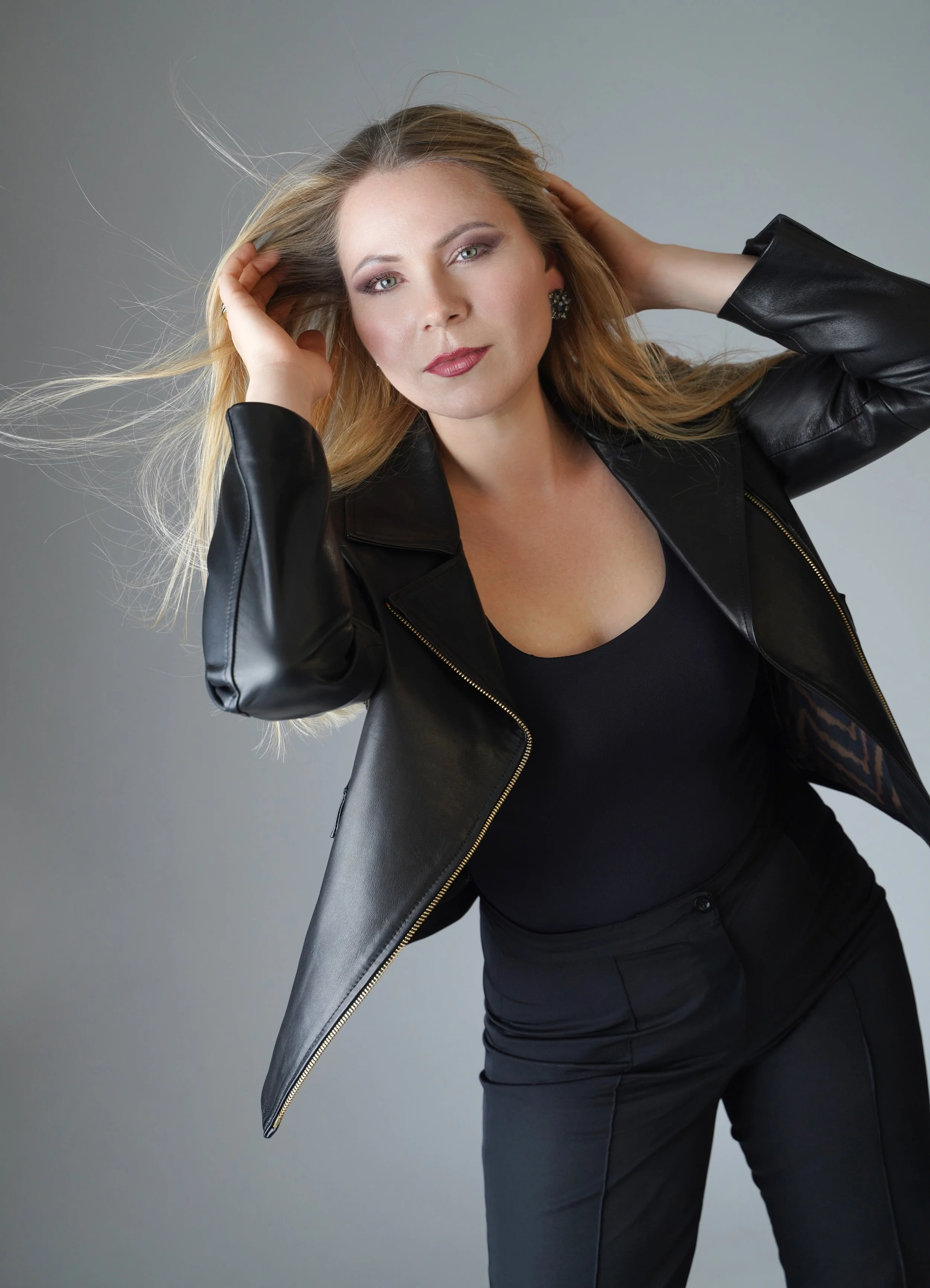 A woman with blonde hair, wearing makeup, earrings, a black top, and a black leather jacket, posing with her hands behind her head against a gray background.