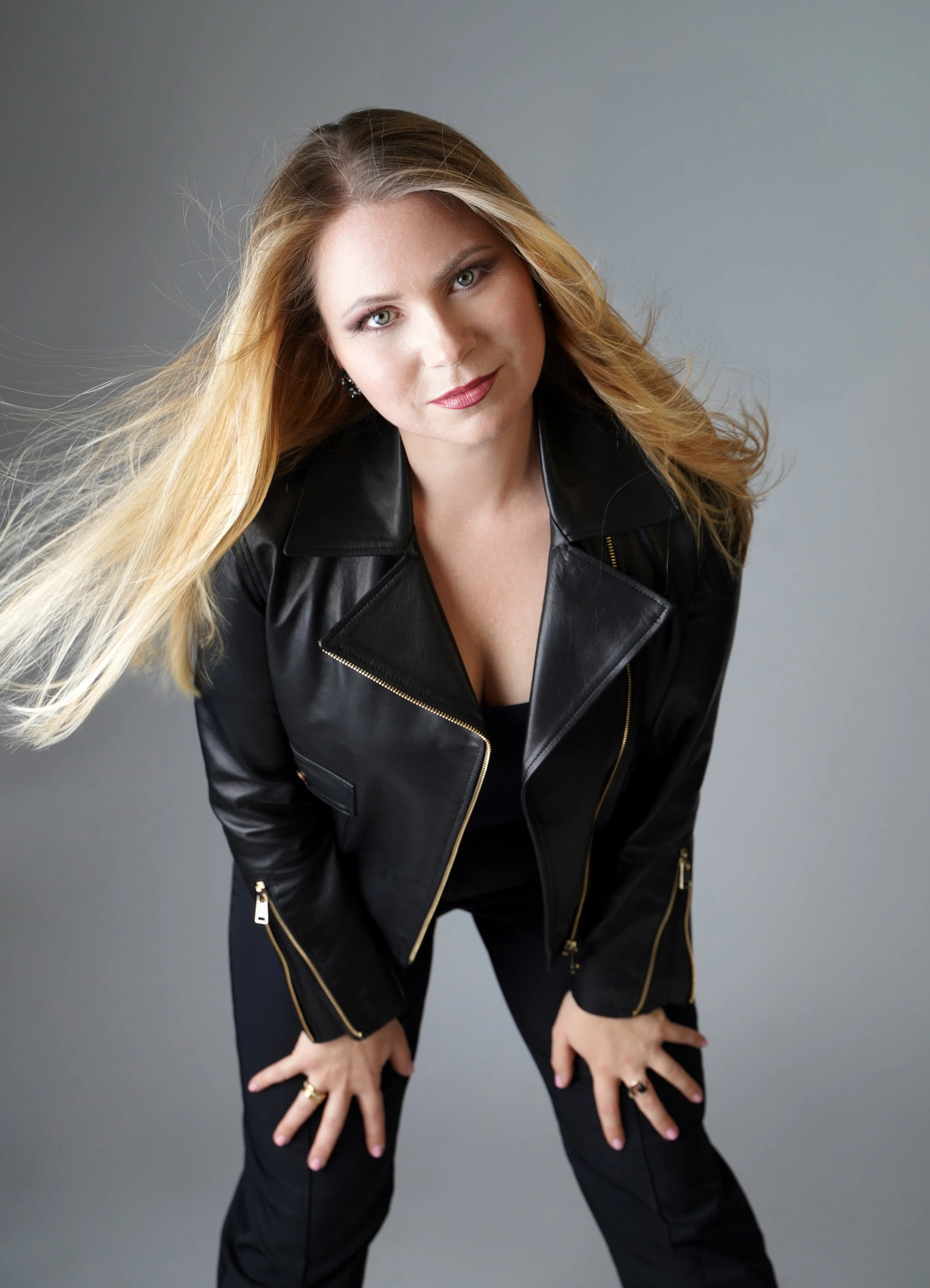 A woman with long blonde hair, wearing a black leather jacket and black pants, is looking at the camera with a confident expression against a plain gray background.
