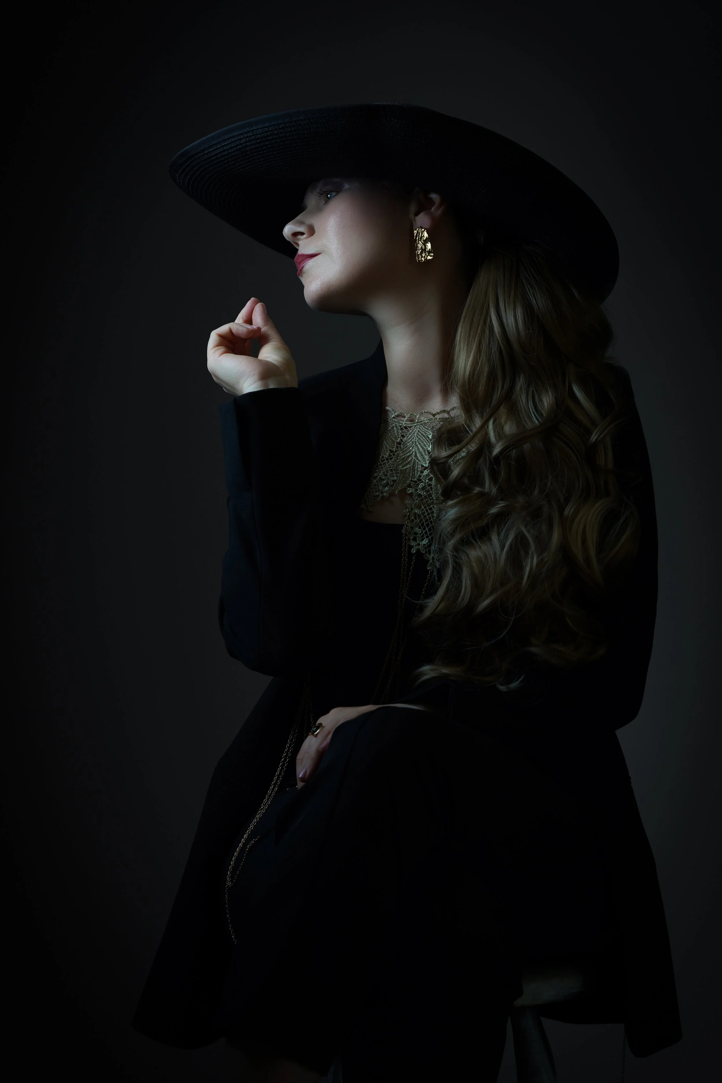A woman with long, curly hair, wearing a wide-brimmed black hat, gold earrings, a dark blazer, and a lace top, is posing against a plain dark background. She is looking to her left with her hand near her face.