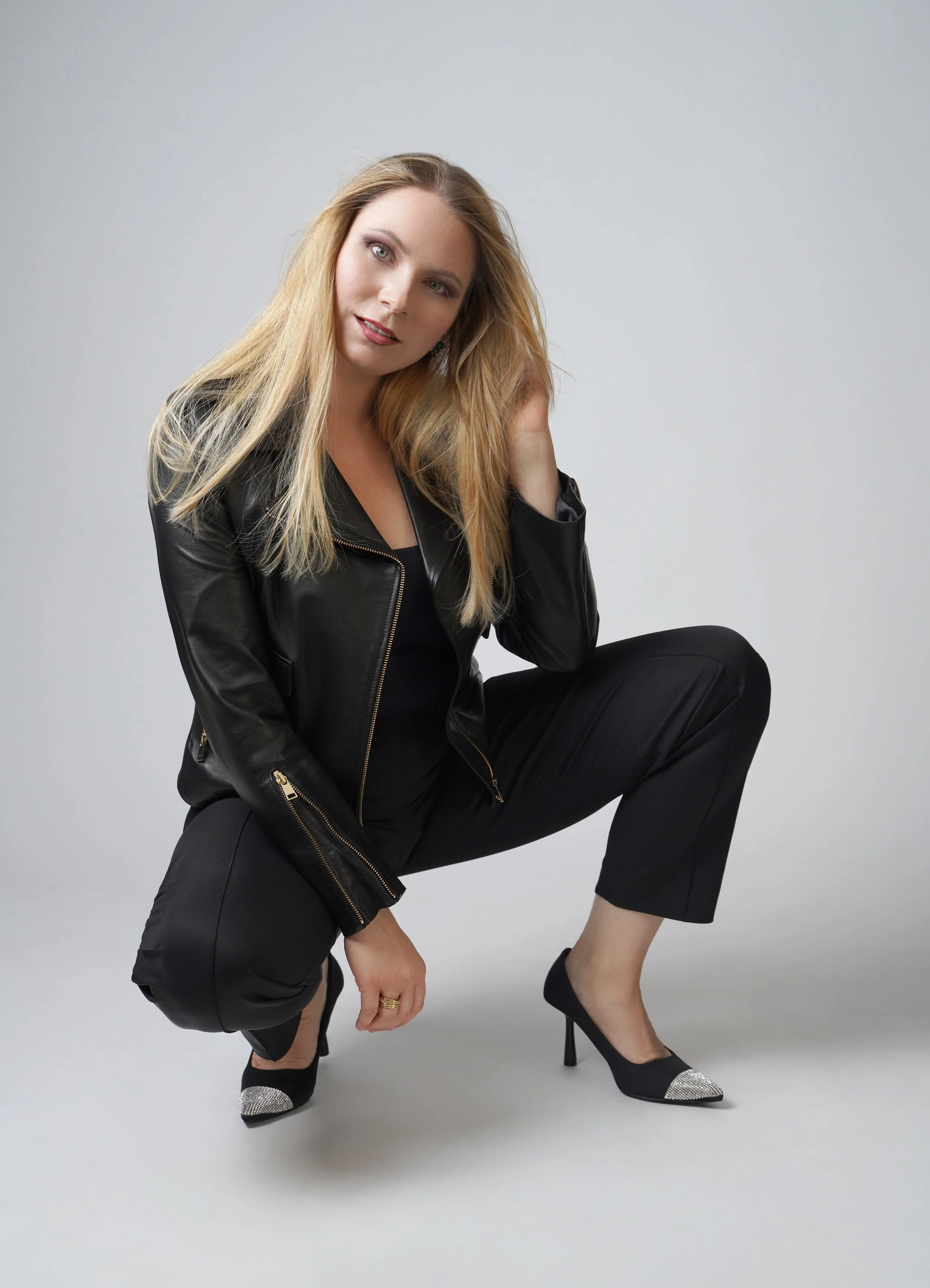 A woman with long blonde hair, wearing a black leather jacket, black pants, and black high heels with glittery tips, posing against a plain white background. She is squatting, with her left hand on her head and her right hand near her knee, looking a