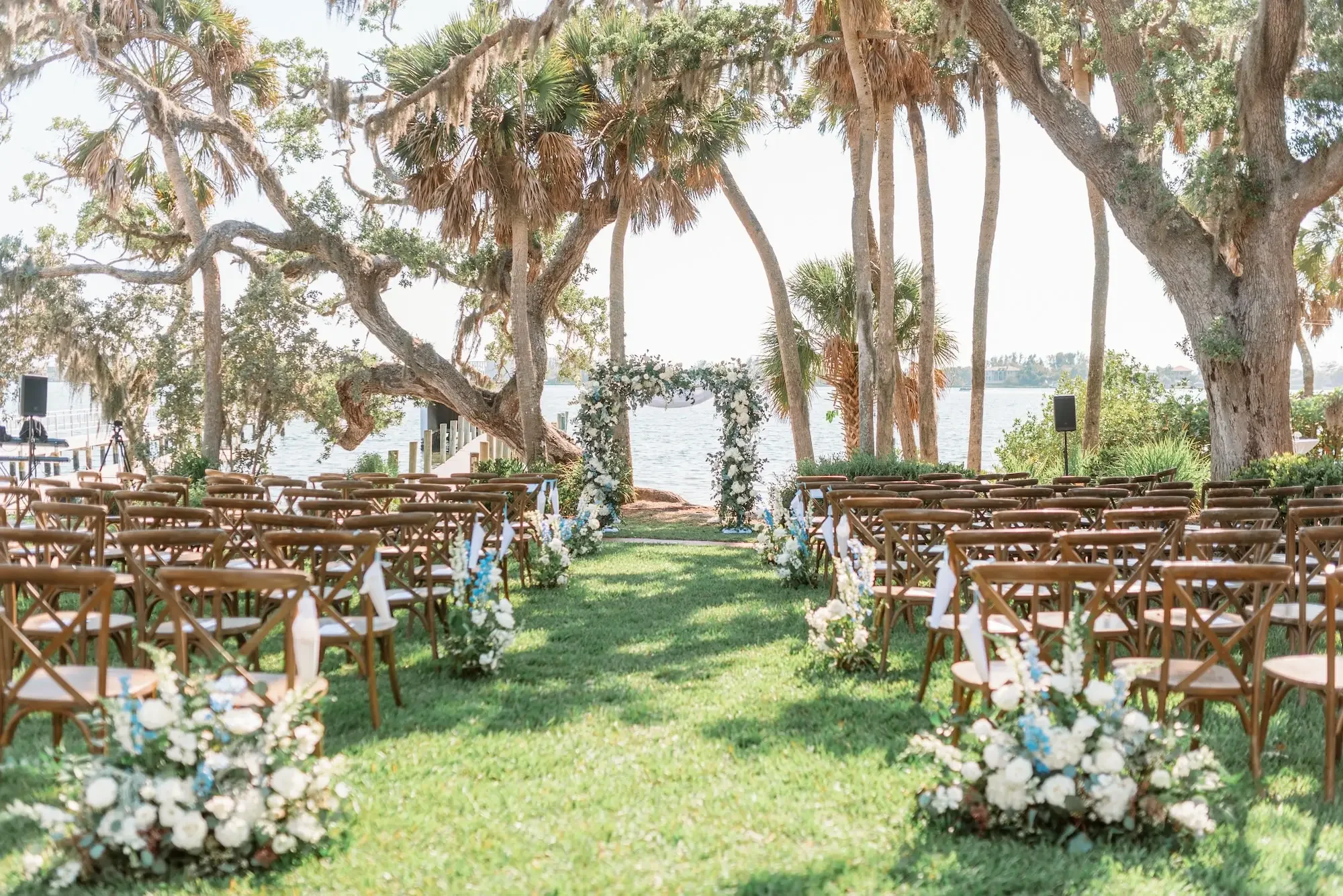 Outdoor wedding ceremony setup with rows of wooden chairs facing an arch decorated with white and blue flowers, surrounded by large trees near a body of water.