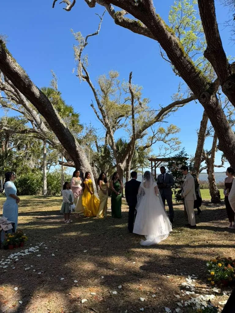 A wedding ceremony taking place outdoors under large trees with sparse leaves. The bride in a white gown and veil is standing with the groom in a dark suit. Several bridesmaids in colorful dresses and groomsmen in suits are present, with some guests standing nearby.