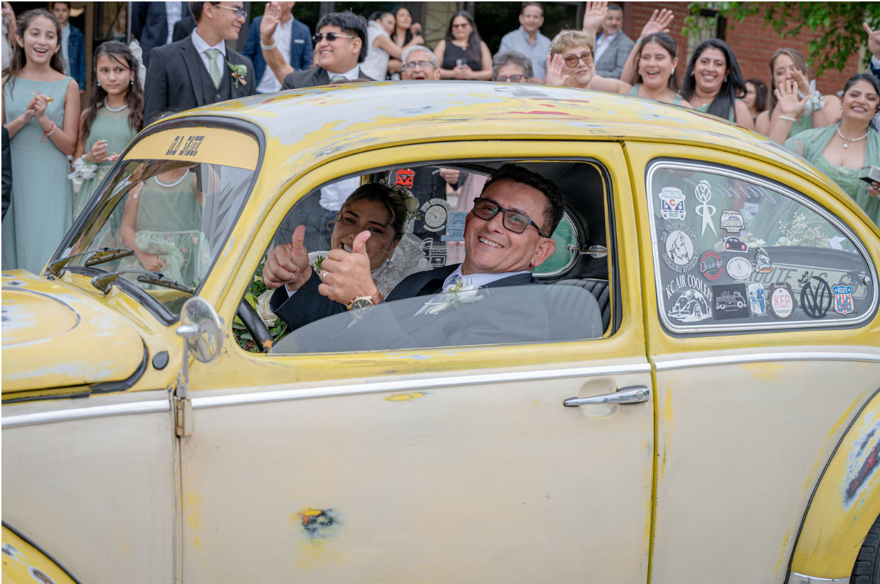 A man and woman sitting in a yellow vintage car decorated for a wedding, surrounded by a crowd of smiling wedding guests, with the man giving a thumbs-up.