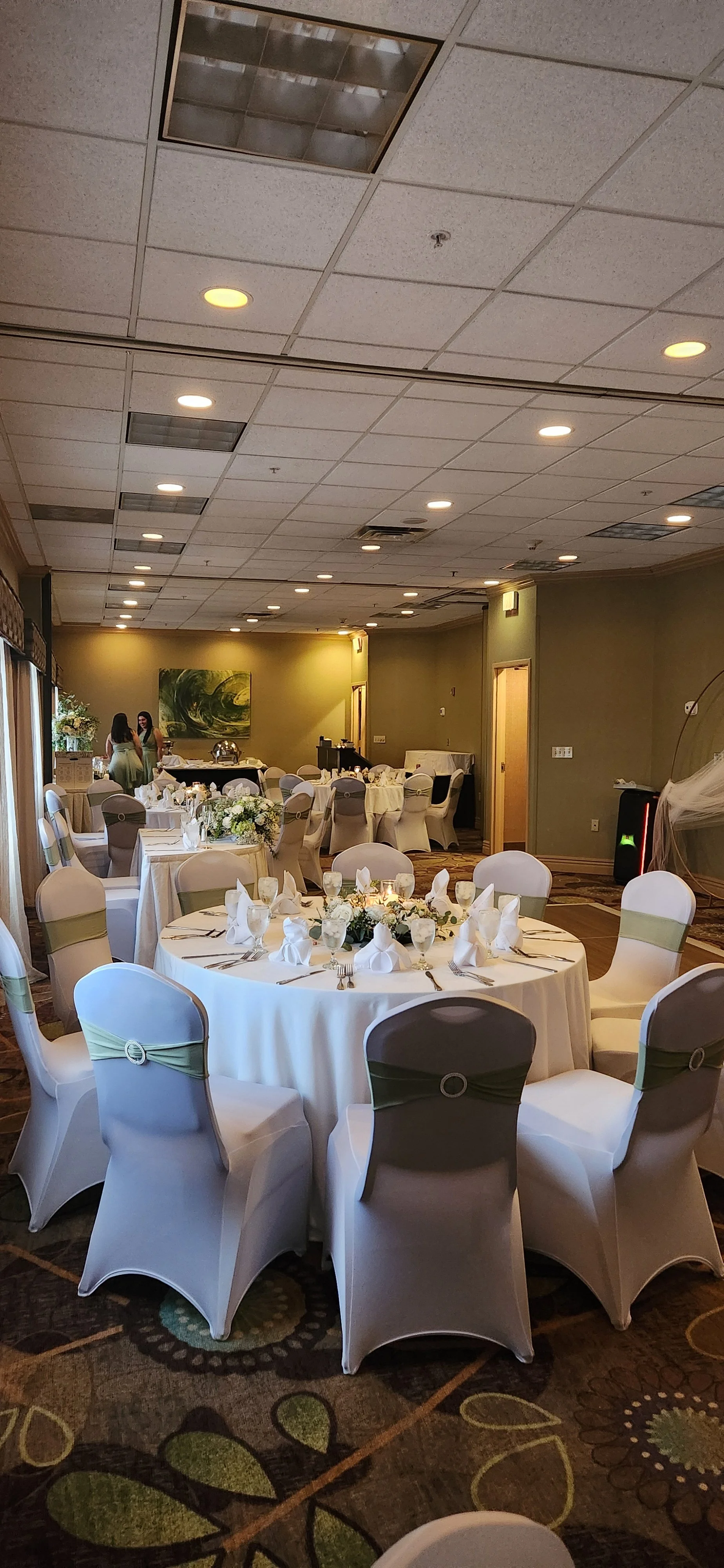 A banquet hall decorated for a wedding reception with round tables covered in white tablecloths, elegant set diningware, and floral centerpieces. The chandeliers and green walls add to the sophisticated ambiance.