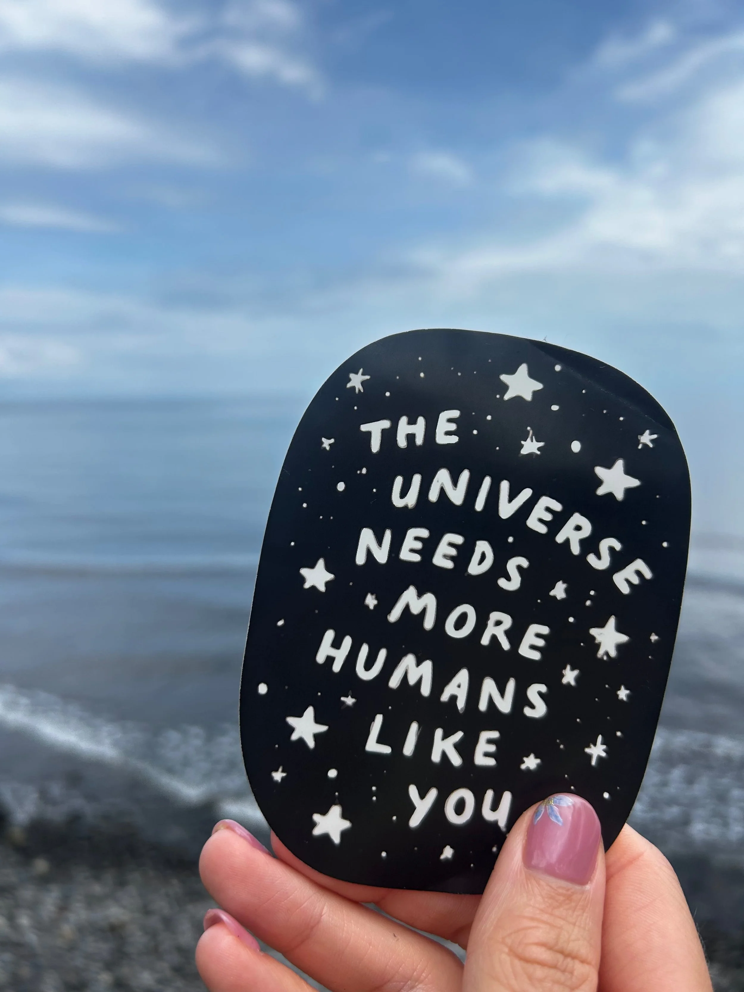 Eine Hand hält ein schwarzes Schild mit weißer Aufschrift: „The universe needs more humans like you“, verziert mit Sternen und Punkten. Im Hintergrund sind ein wolkiger Himmel und das Meer zu sehen.