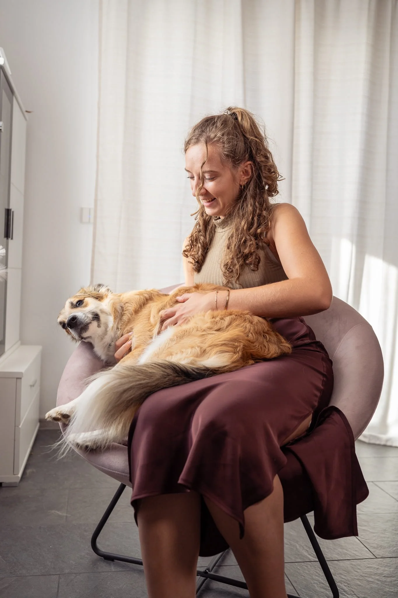 Eine Frau mit lockigem Haar sitzt lächelnd auf einem rosafarbenen Stuhl, streichelt und spielt mit einem hellem Mischlingshund, der auf ihrem Schoß liegt. Die Szene befindet sich in einem Raum mit weißen Wänden und Vorhängen.
