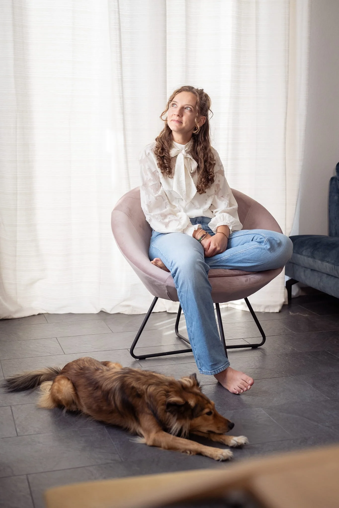 Eine junge Frau mit lockigem Haar und einer weißen Bluse mit Schleife sitzt mit überschlagenen Beinen auf einem rosafarbenen Stuhl und blickt nachdenklich zur Seite. Vor ihr liegt ein Hund auf dem dunklen Fliesenboden. Im Hintergrund Vorhänge.