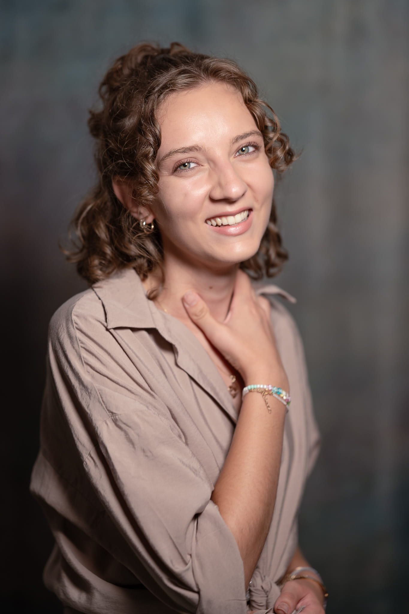 Eine lächelnde Frau mit lockigem braunem Haar, blauen Augen und heller Haut berührt mit ihrer rechten Hand ihren Hals. Sie trägt ein beiges Oberteil und bunte Armbänder.