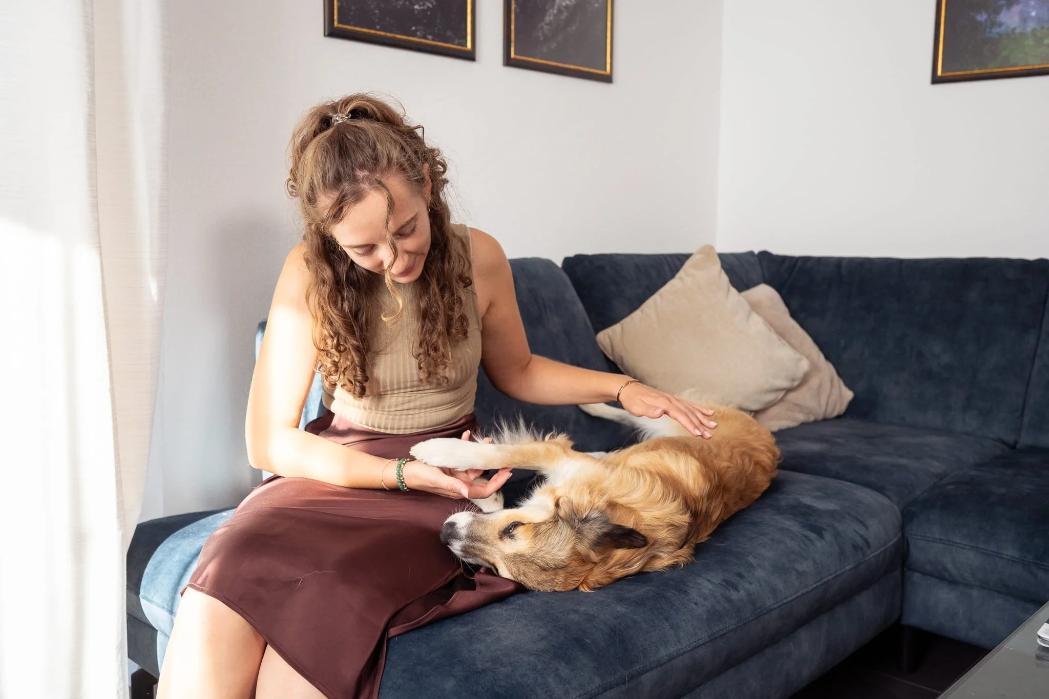 Eine Frau mit langen, lockigen Haaren sitzt auf einem dunkelblauen Sofa und macht sanft Energiearbeit bei einem entspannt auf dem Rücken liegenden Hund. Sie trägt ein beigefarbenes Oberteil und einen roten Rock.