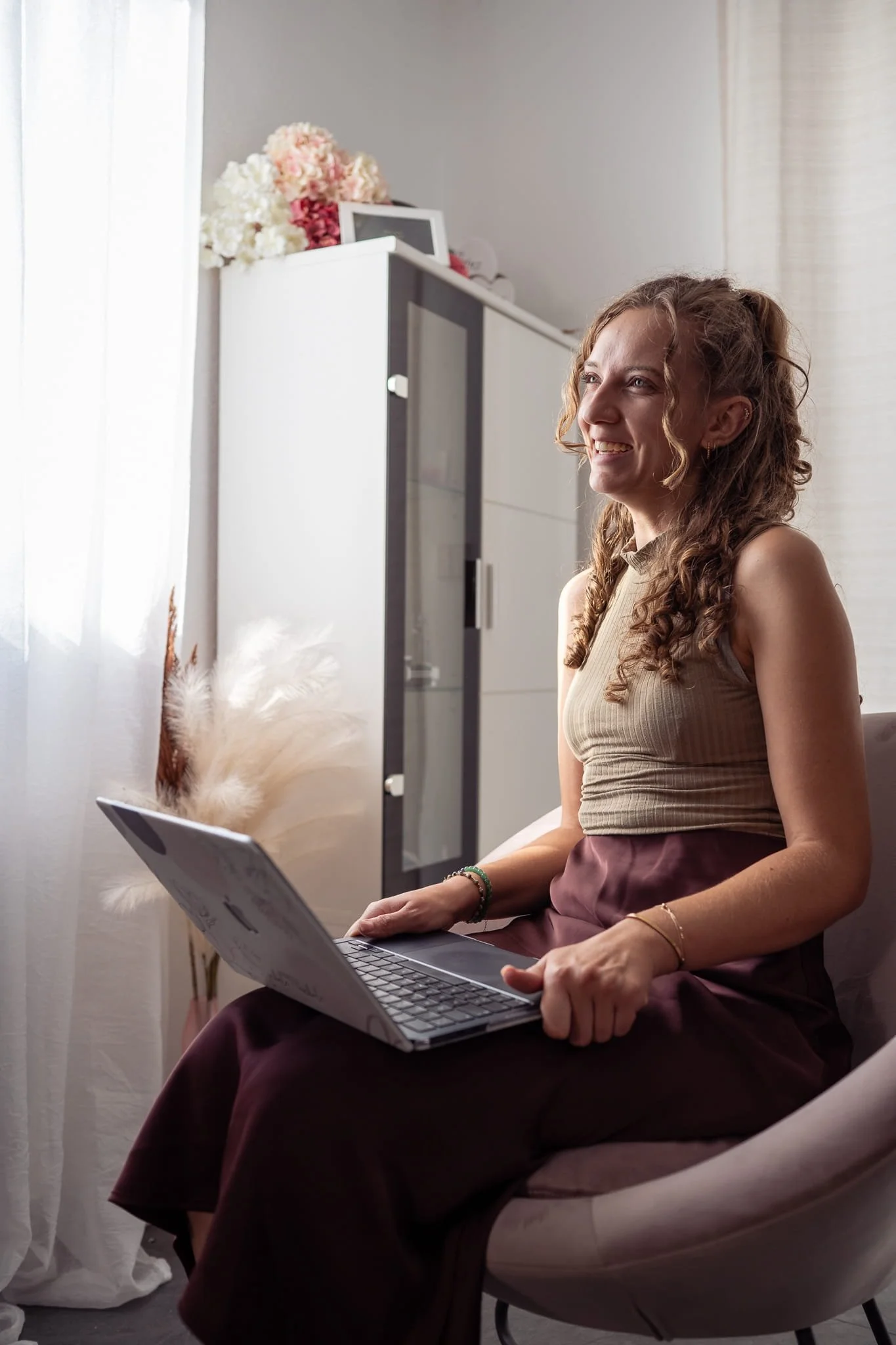 Eine Frau mit lockigem Haar lächelt, während sie auf einem modernen Stuhl sitzt und an einem Laptop arbeitet. Sie befindet sich in einem hellen Raum mit Vorhängen und dekorativen Blumen im Hintergrund.