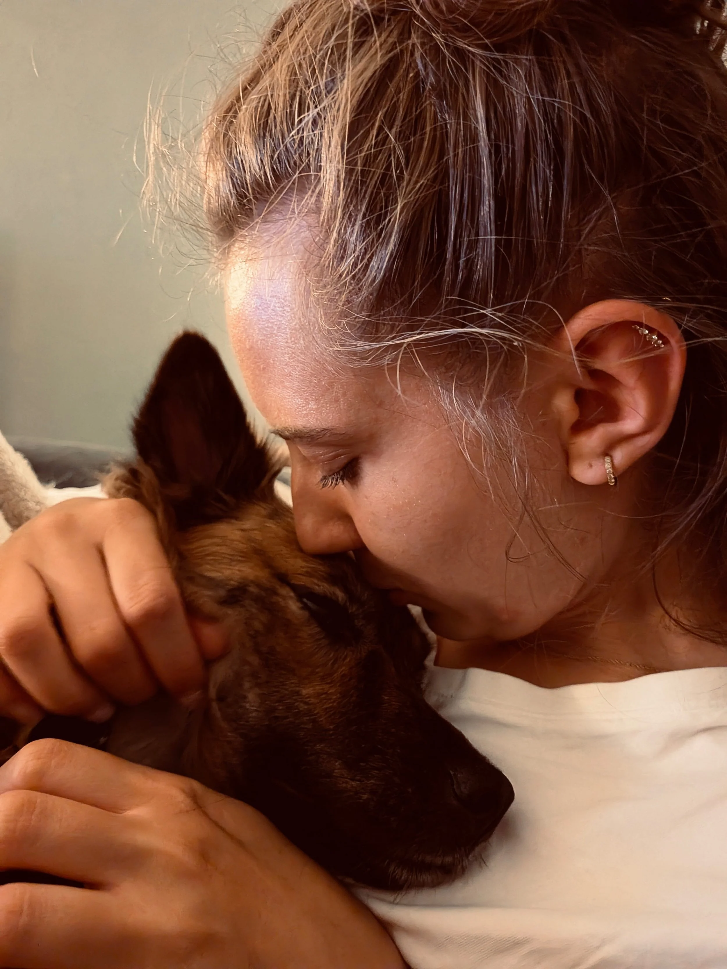 Eine Person mit lockigen braunen Haaren und gepiercten Ohren, die einen kleinen braunen Hund mit spitzen Ohren und geschlossenen Augen hält.