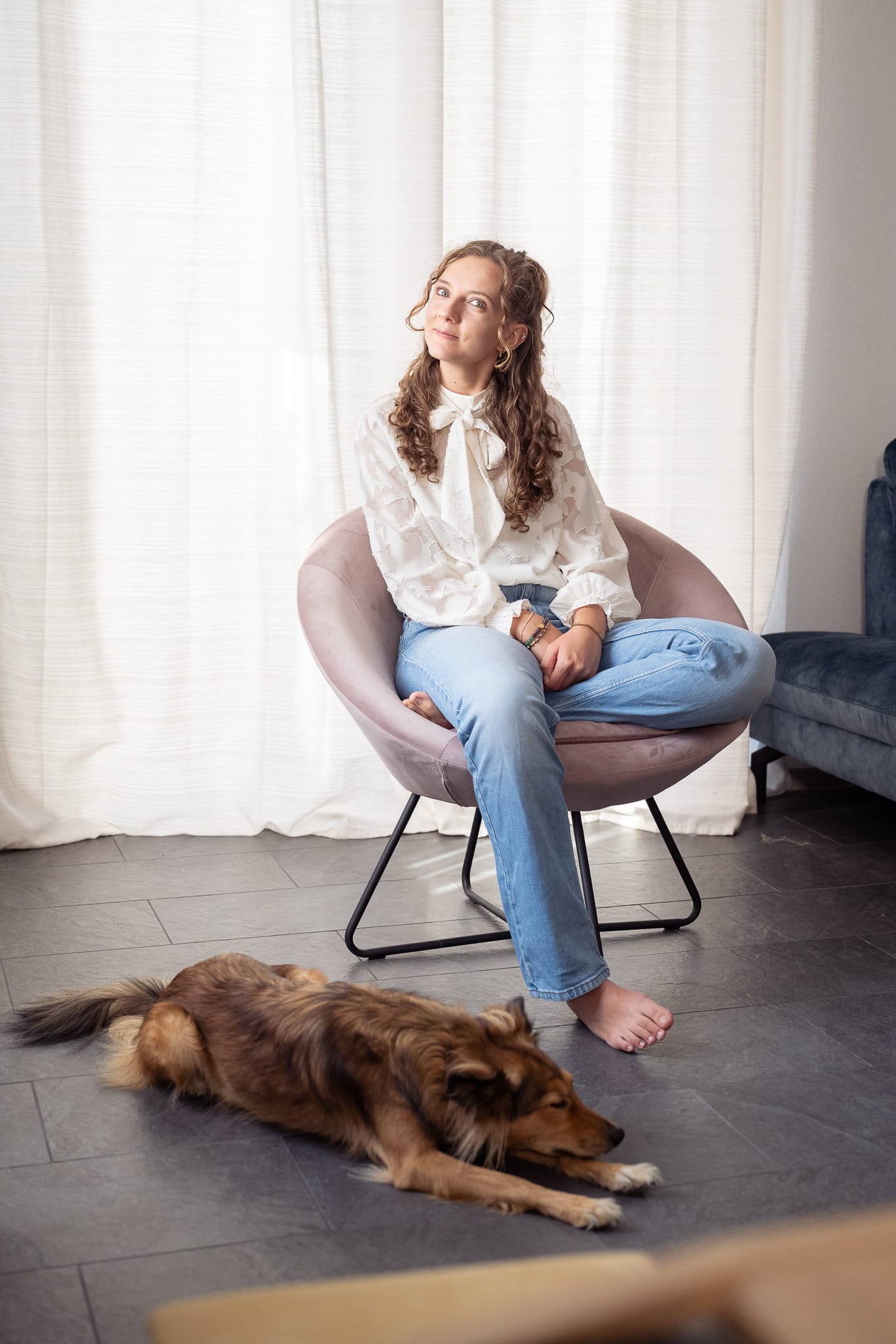 Eine Frau mit lockigem Haar sitzt auf einem rosafarbenen Stuhl und trägt eine weiße Bluse und hellblaue Jeans. Neben ihr liegt ein Hund auf dem Boden.