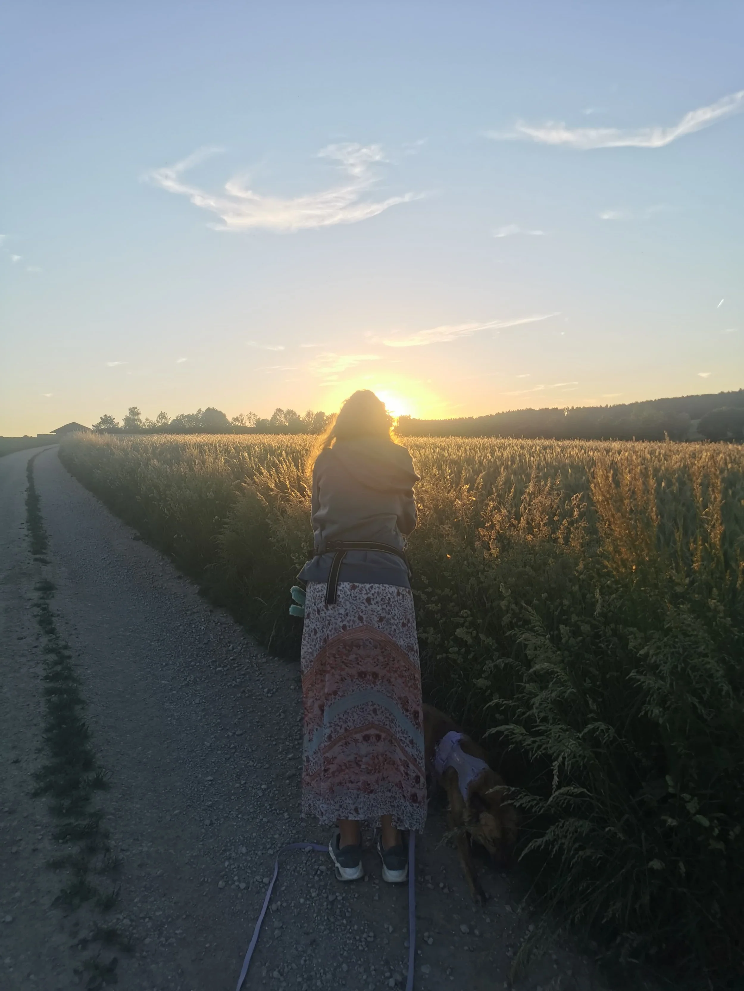 Eine Frau steht auf einem Schotterweg und blickt in Richtung Sonne. Dabei fotografiert leicht nach vorne gebeugt den Sonnenuntergang über dem Feld. Sie trägt Sneaker und einen gemustertes Kleid.