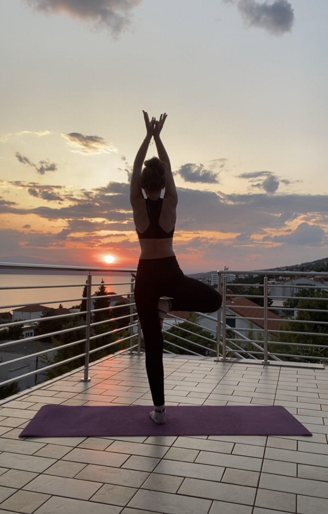 Eine Person, die bei Sonnenuntergang auf einem Balkon Yoga praktiziert. Im Hintergrund sind der Sonnenuntergang, der Horizont und das Meer zu sehen.