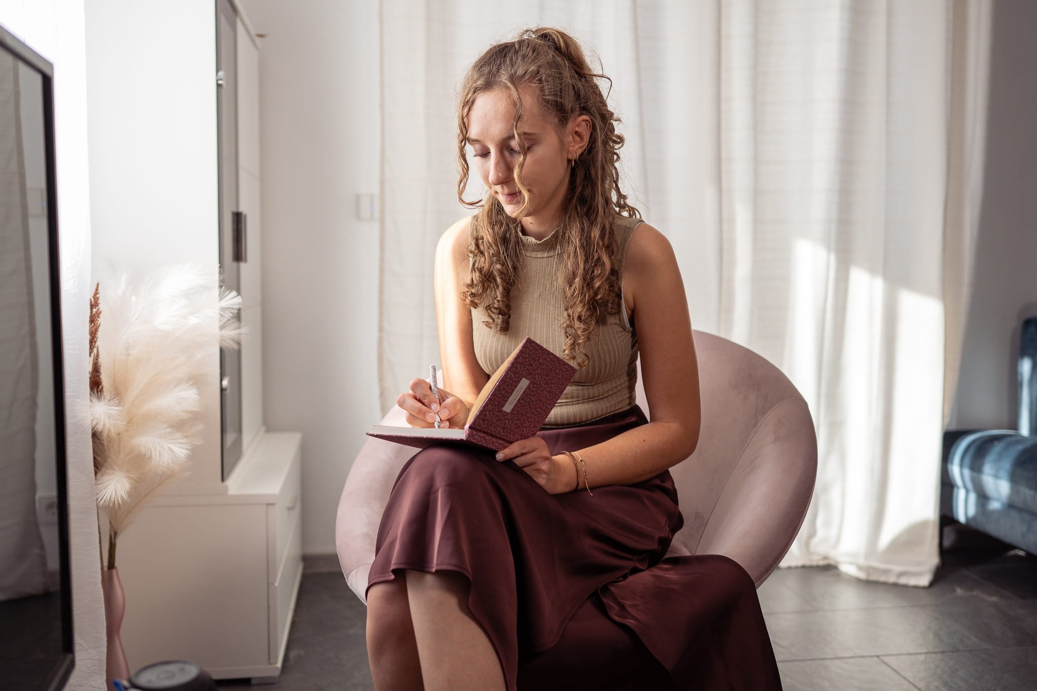 Eine junge Frau mit lockigem Haar sitzt auf einem rosafarbenen Stuhl und schreibt in ein Notizbuch. Sie trägt ein ärmelloses beigefarbenes Oberteil und einen roten Rock. Die Szene spielt in einem modernen, hellen Raum mit Vorhängen.