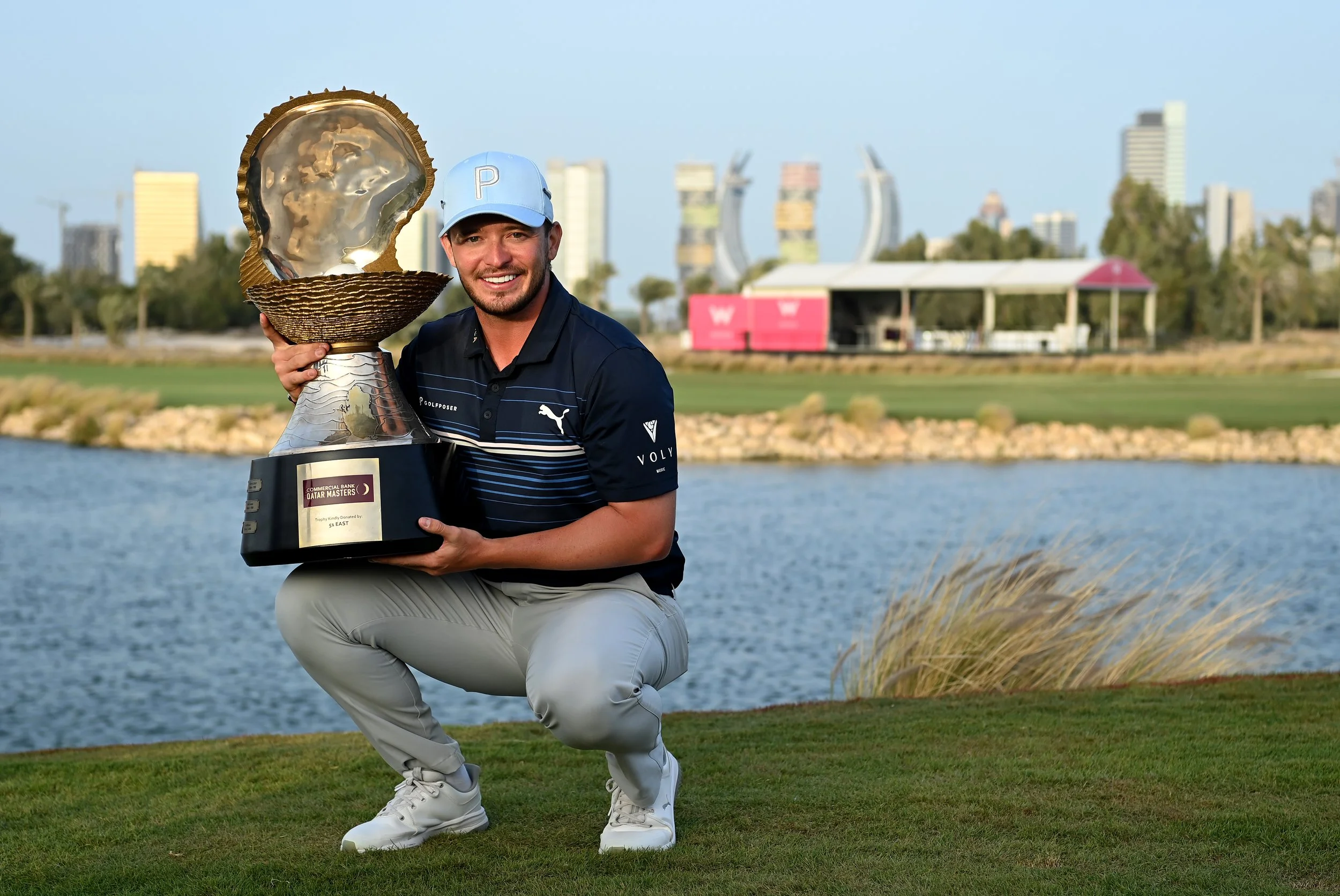 Man kneeling on a golf course holding a large trophy with a shell-shaped top. He is wearing a blue cap, black polo shirt, and light-colored pants. The background features a body of water, trees, and city buildings.