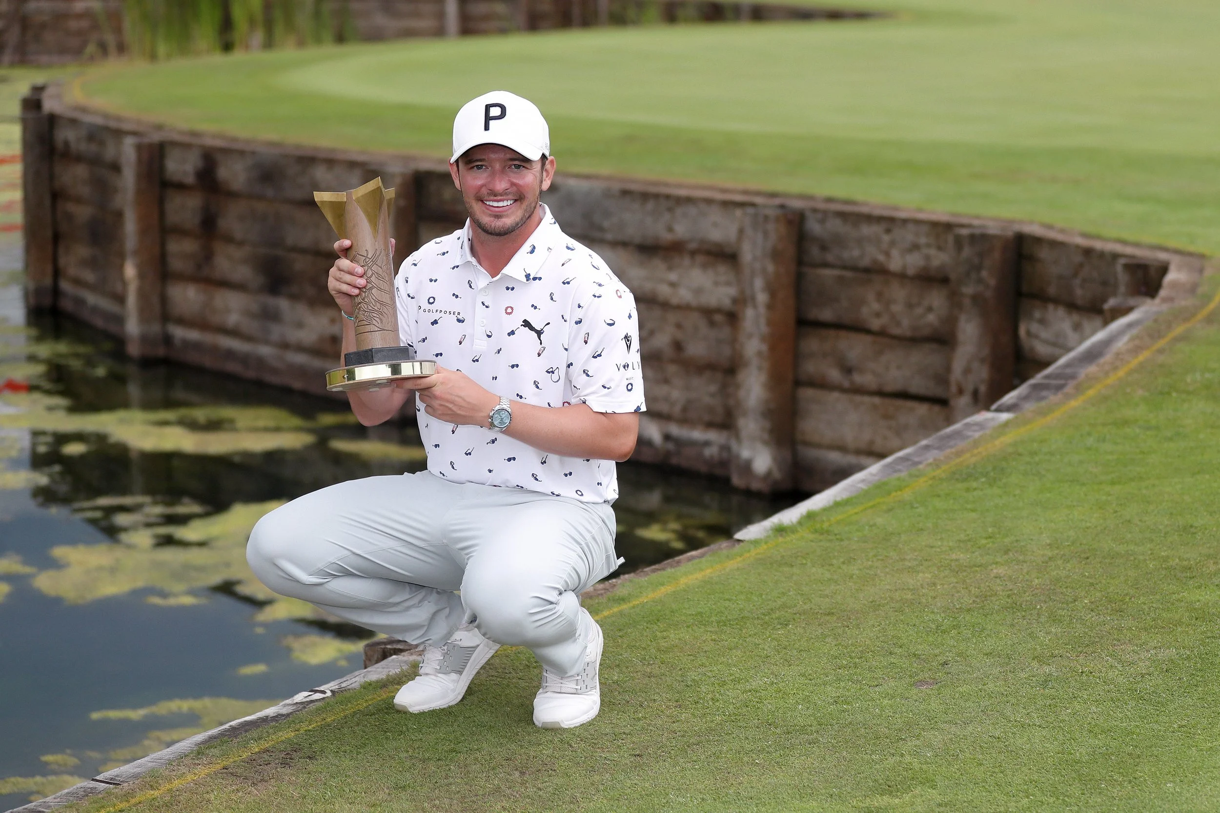 A man kneeling on a golf course holding a trophy, smiling, with a green field, water, and a wooden fence behind him.