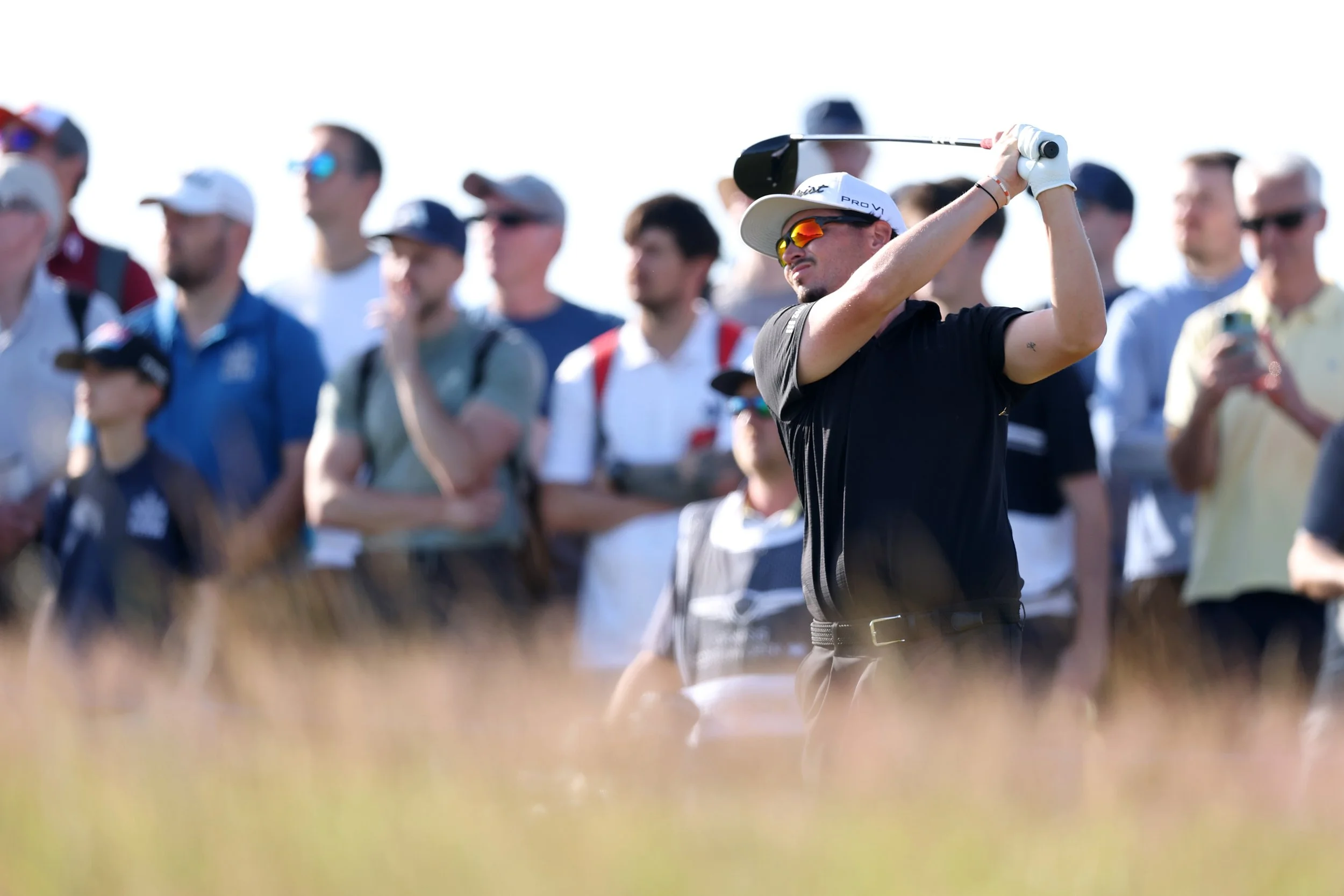 A man in a black shirt and white cap, wearing sunglasses, is swinging a golf club. He is surrounded by a group of people, some wearing hats and sunglasses, on what appears to be a golf course on a sunny day.