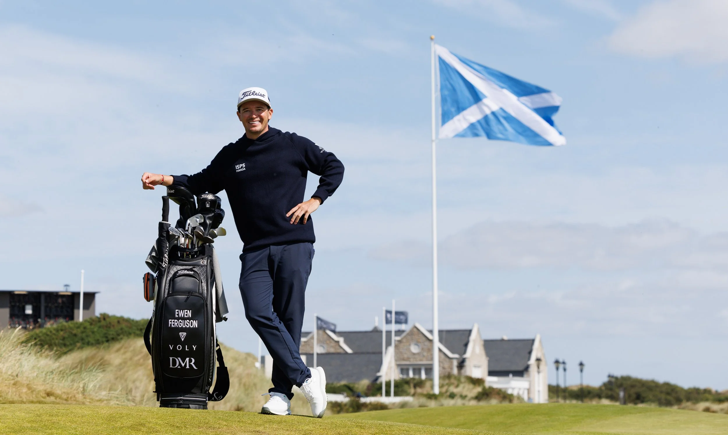 A male golfer standing on a golf course, smiling, with a golf bag and clubs. There is a blue and white flag and houses in the background.