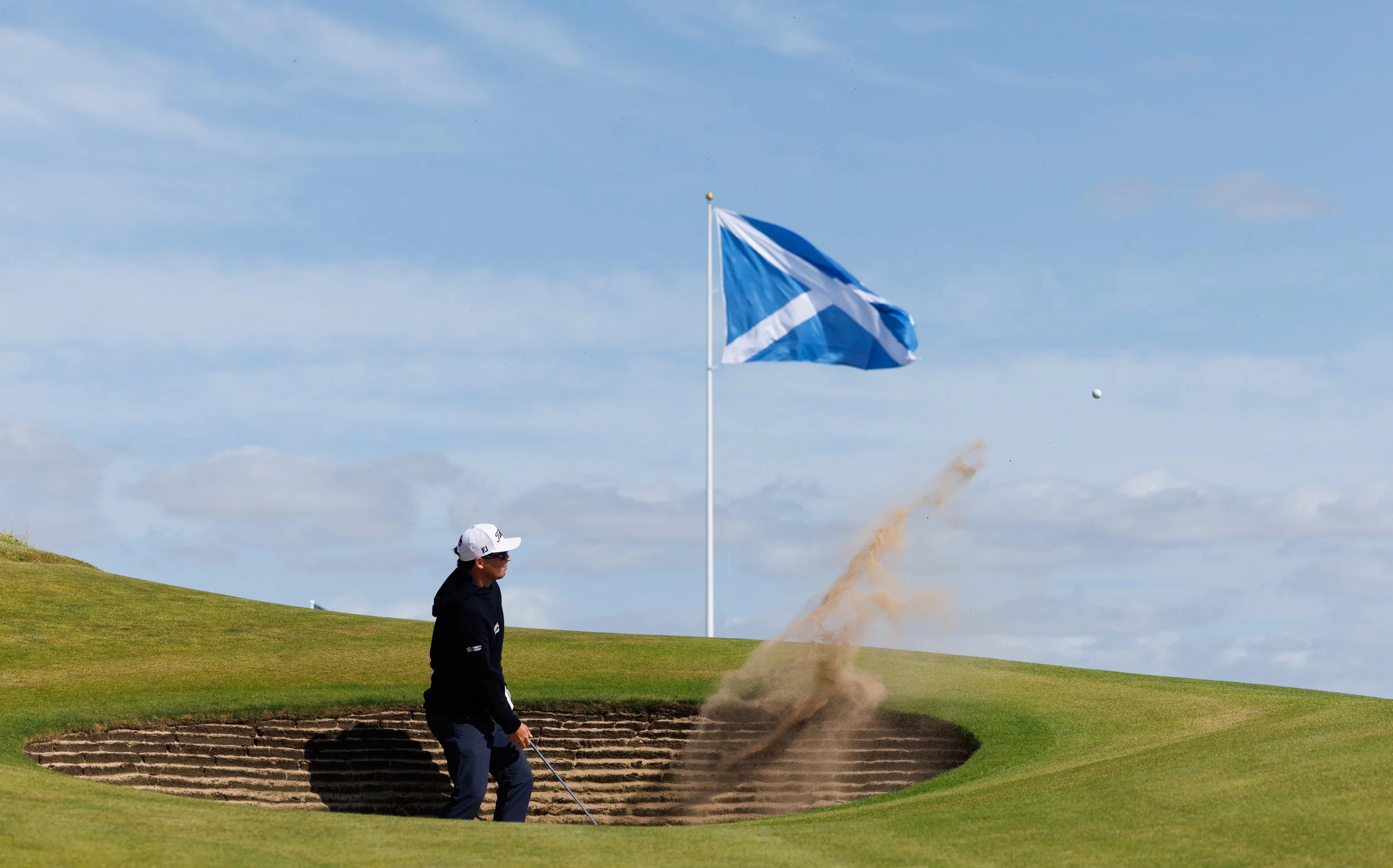 A golfer on a golf course preparing to putt, with a blue and white flag in the background, a sand bunker, and a blue sky with some clouds.