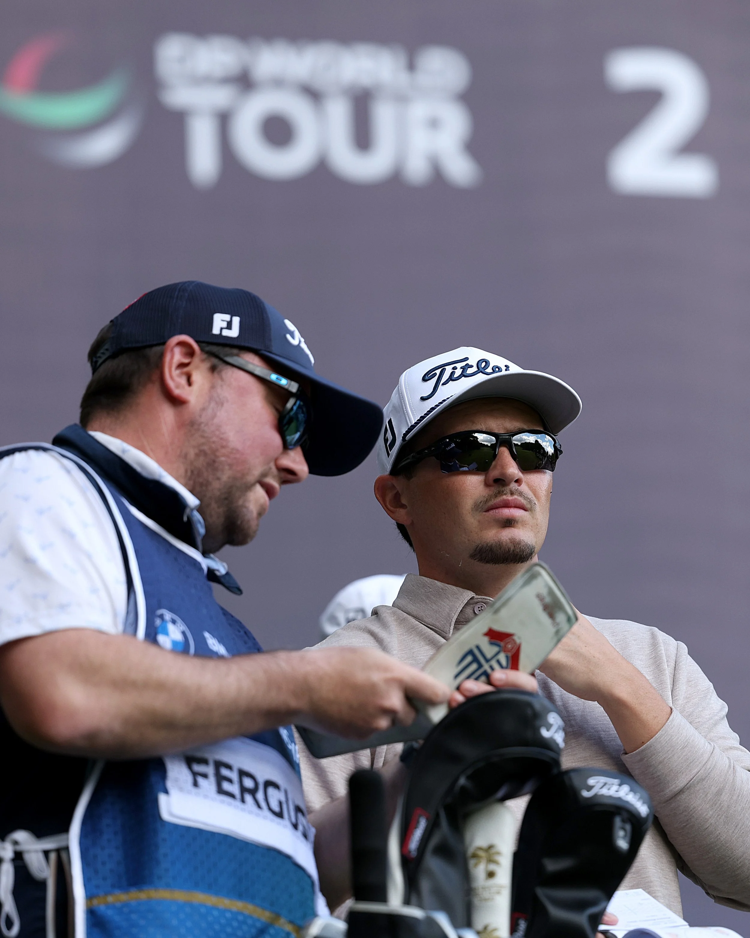 Two professional golfers reviewing golf clubs and a scorecard with sponsor logos, wearing golf caps and sunglasses, at a golf tournament awards ceremony.