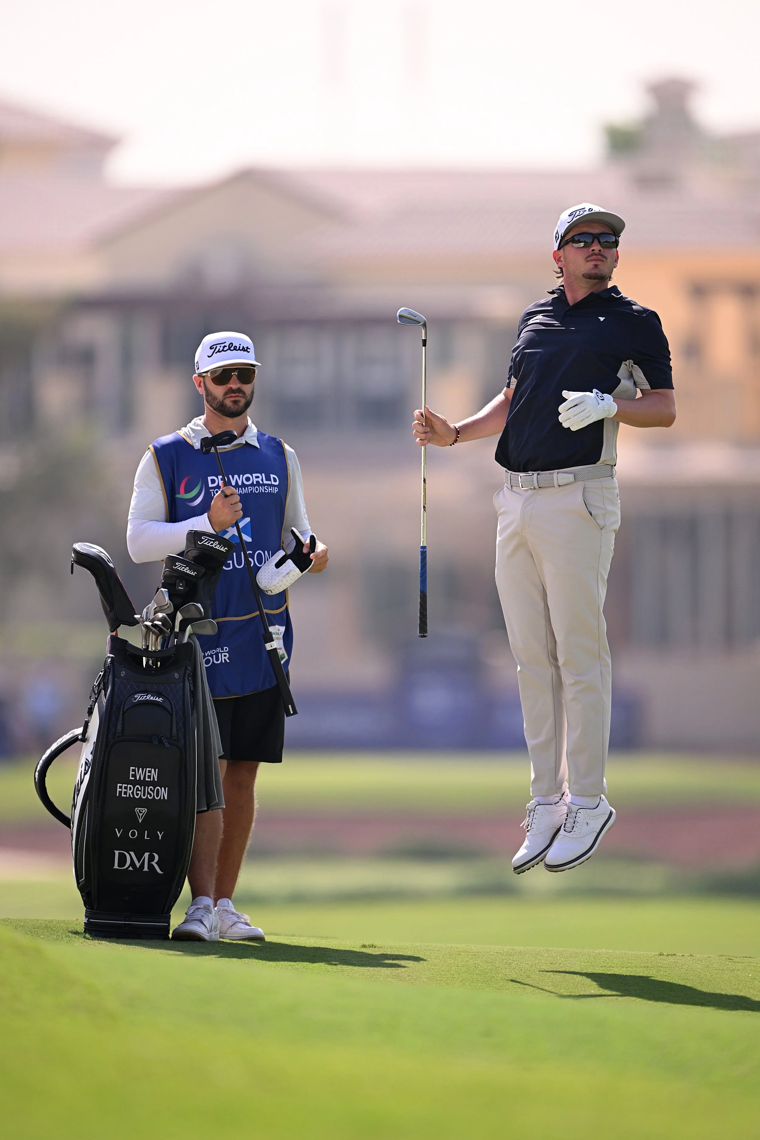 A golfer is mid-air after hitting a shot on a golf course, with another caddie standing nearby.