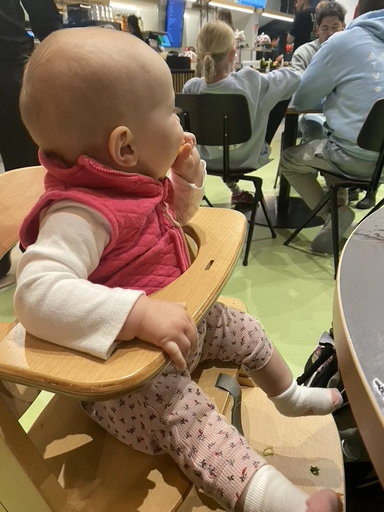 A young child with a bald head sitting in a high chair at a food court or cafe, wearing a pink vest, white long-sleeve shirt, and patterned pants, with one hand near their mouth and the other resting on the high chair.