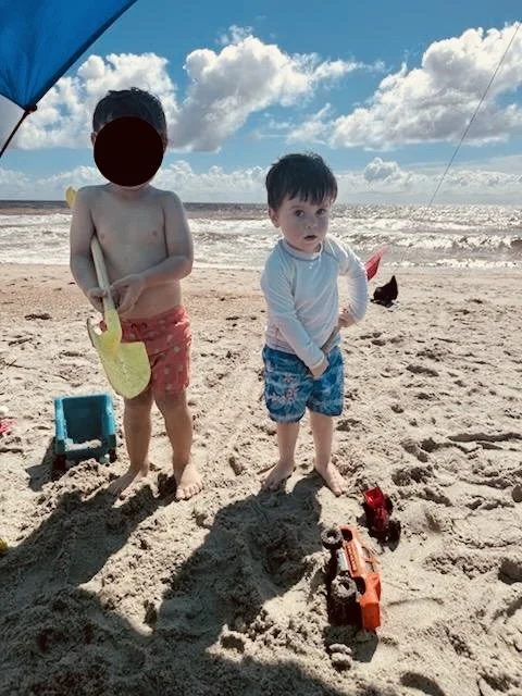 Two young boys playing on the sandy beach with toys, one holding a green shovel and the other with a red toy truck, under a blue beach umbrella with the ocean and sky with clouds in the background.