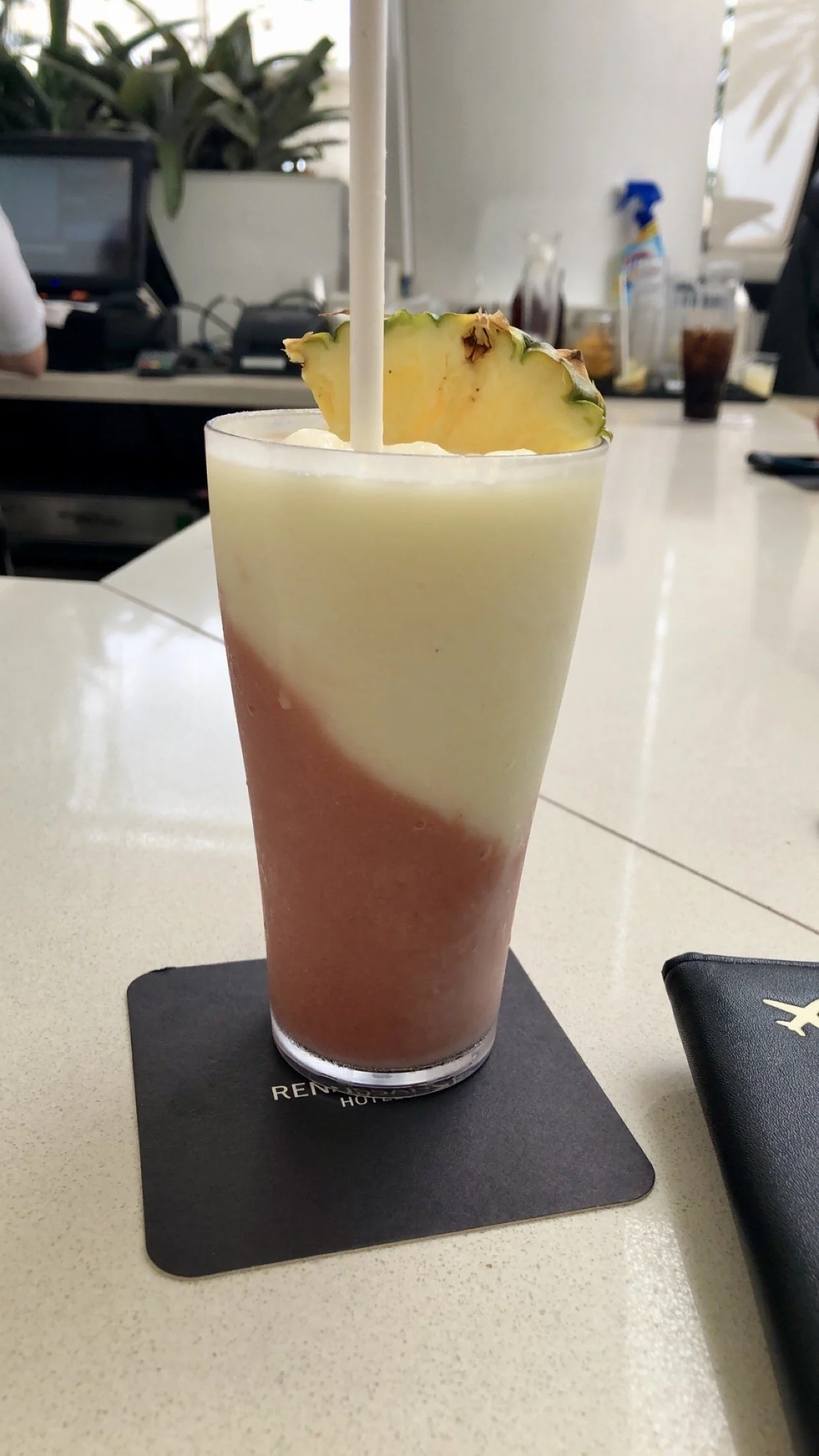 A multi-layered iced drink with a pineapple slice on top, placed on a black coaster on a white table inside a cafe or restaurant.