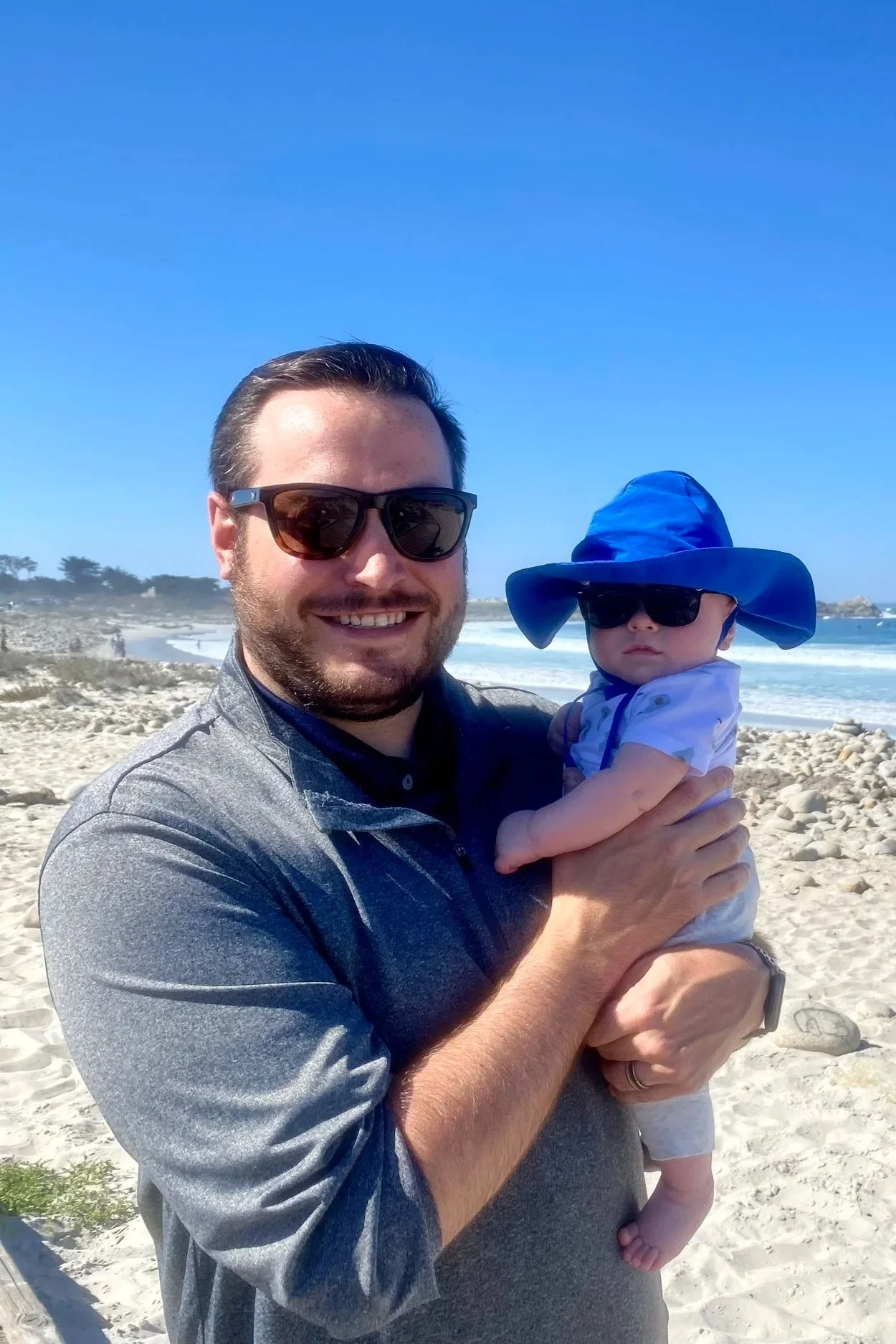 A man with sunglasses smiling and holding a baby on a beach, both wearing sunglasses and the baby wearing a large blue sun hat.