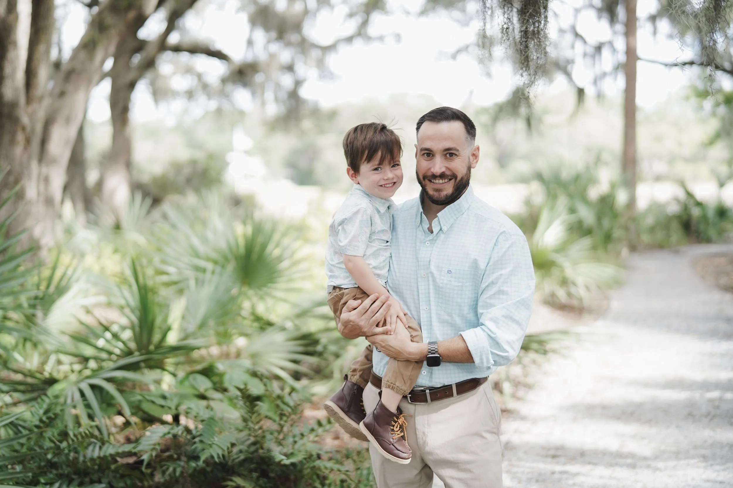 A man holding a young boy outdoors in a lush, wooded area.