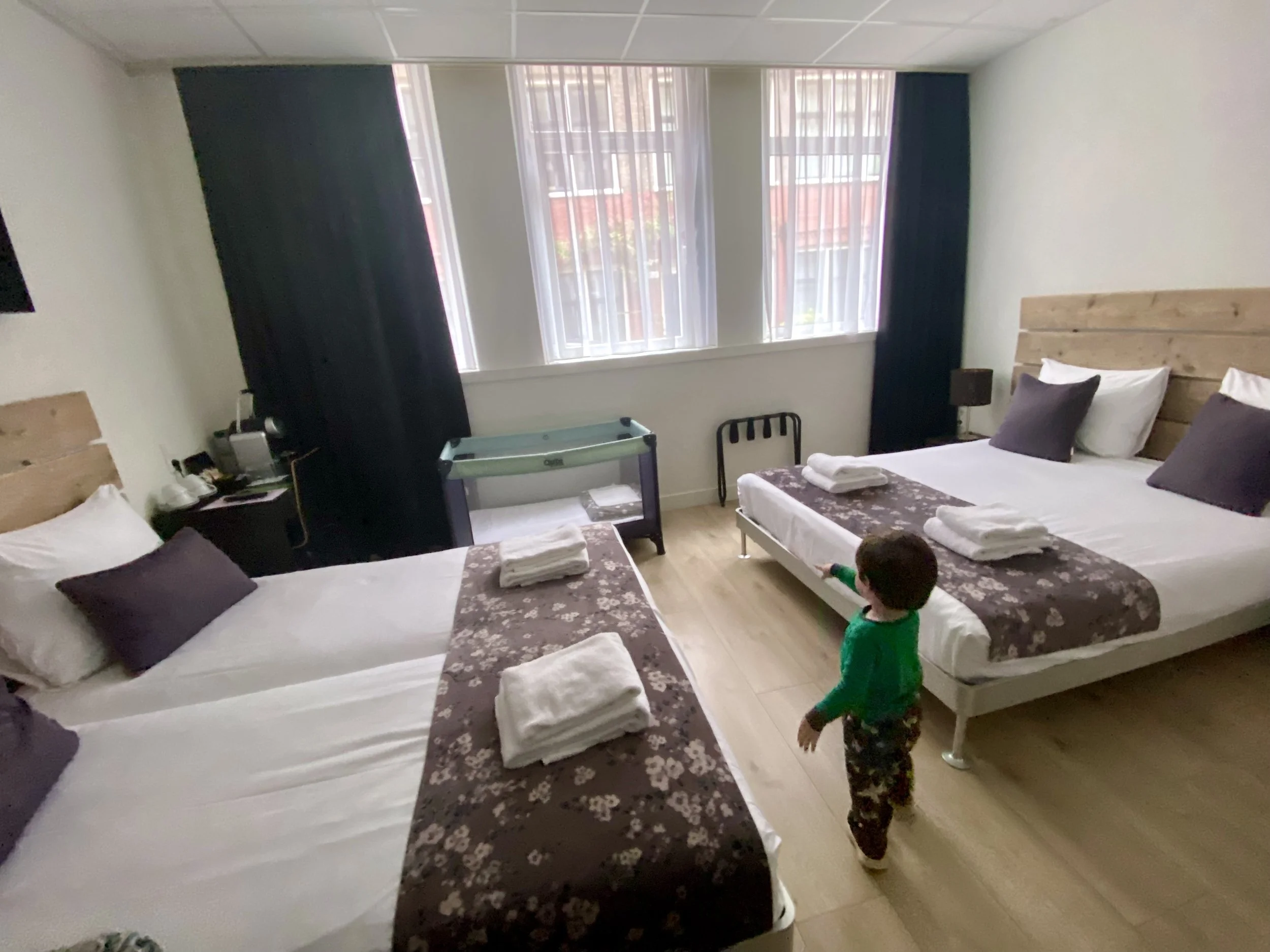 A hotel room with three beds, each made with white sheets, brown floral bed runners, and folded towels on top. There are purple and white pillows on the beds. The room has light-colored wooden flooring, a large window with sheer curtains and blackout drapes, and a small bedside table with a lamp. A young child in a green shirt and patterned pants is walking near the beds.