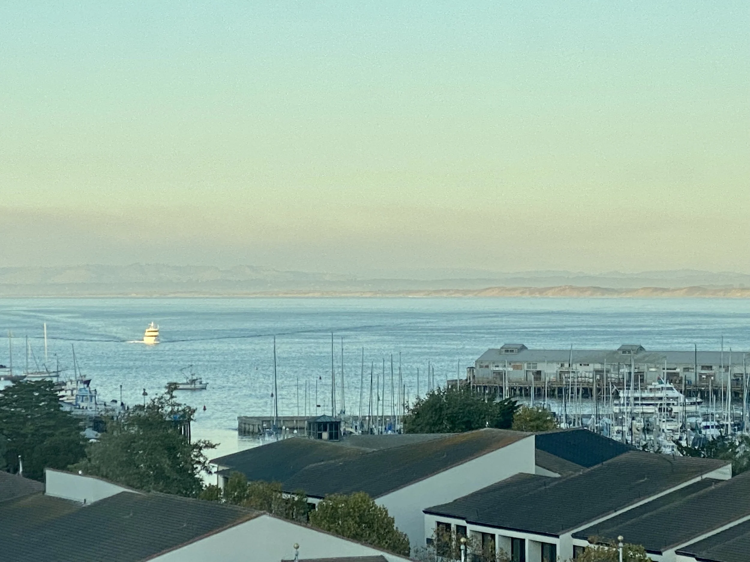 A coastal scene with boats and buildings along the shoreline, view of the ocean, distant land or islands, and a ship sailing on the water during daylight hours.