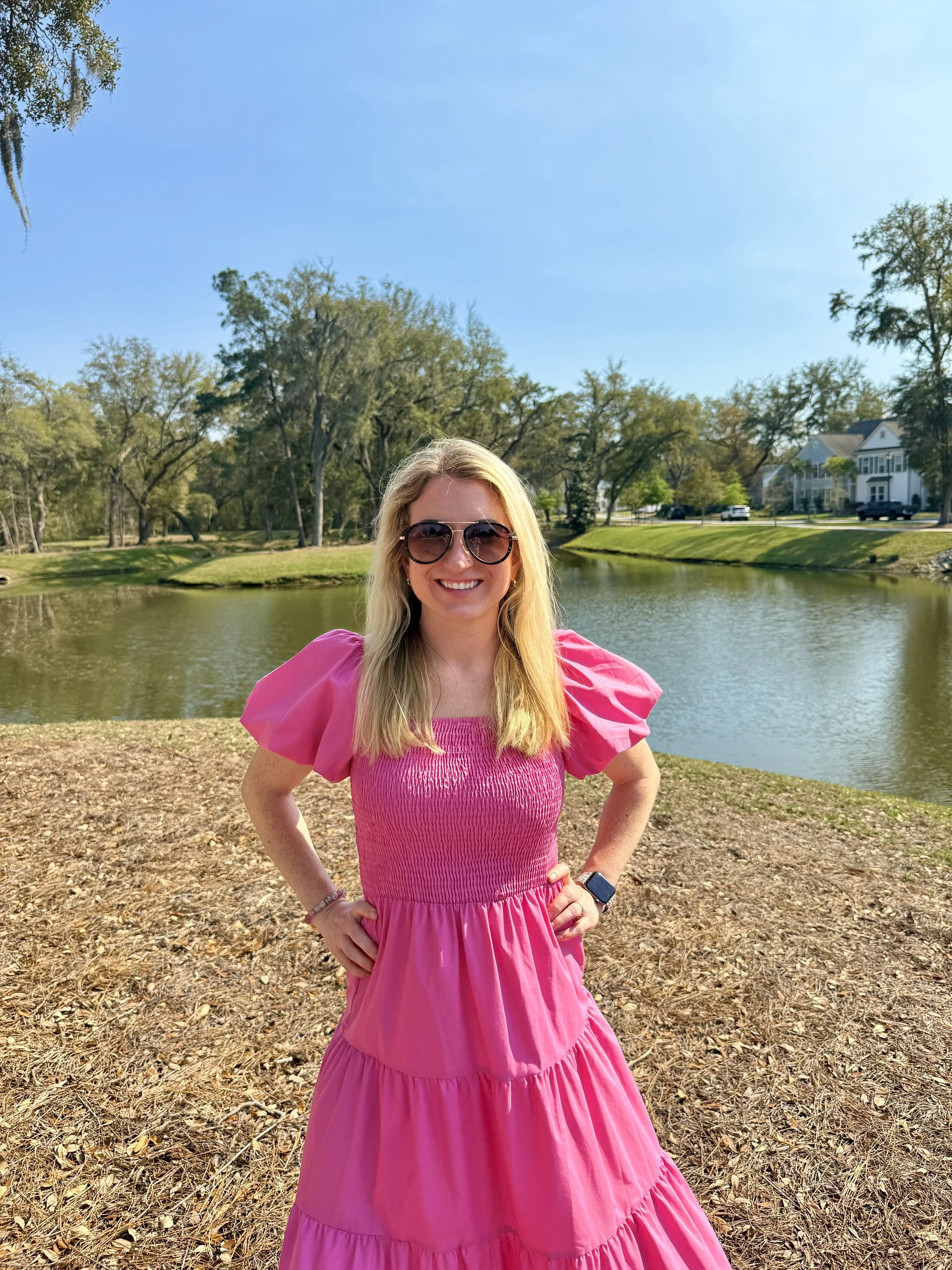 A woman with blonde hair wearing sunglasses and a pink dress, smiling outdoors near a body of water with trees and houses in the background.