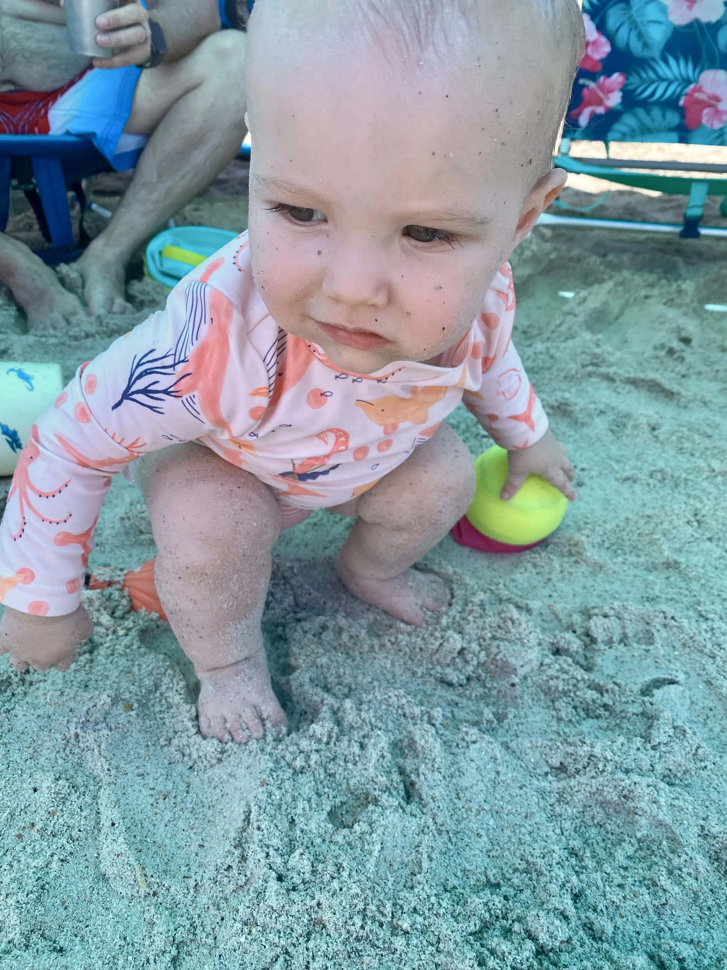 A young child with a shaved head playing in the sand at the beach, holding a yellow ball, surrounded by sand and a few adults in the background.