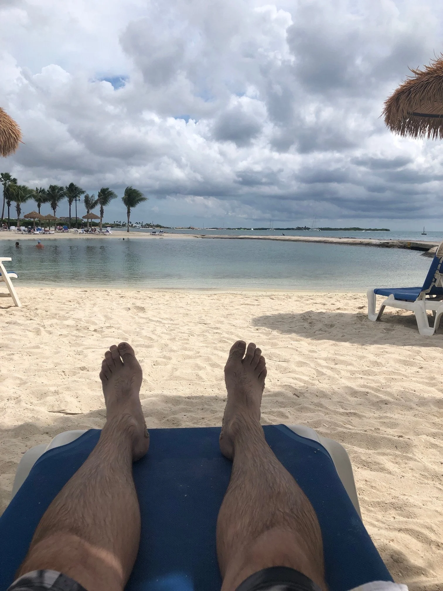 A person relaxing on a lounge chair at the beach, with their legs and feet visible, facing towards the ocean and sandy shoreline, under a cloudy sky.