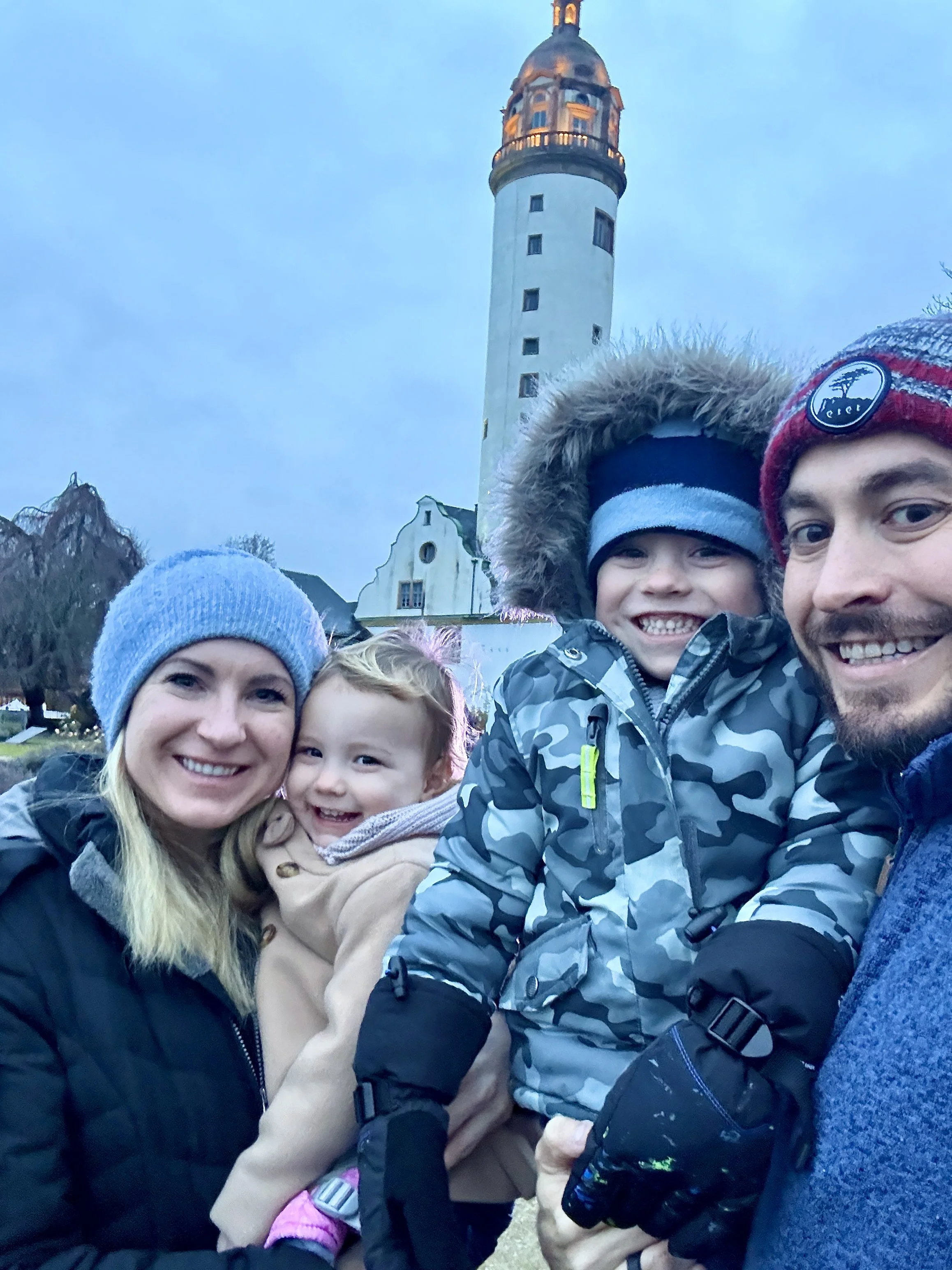 A group of people bundled up in winter clothing posing for a selfie outdoors with a historic lighthouse and a building in the background.