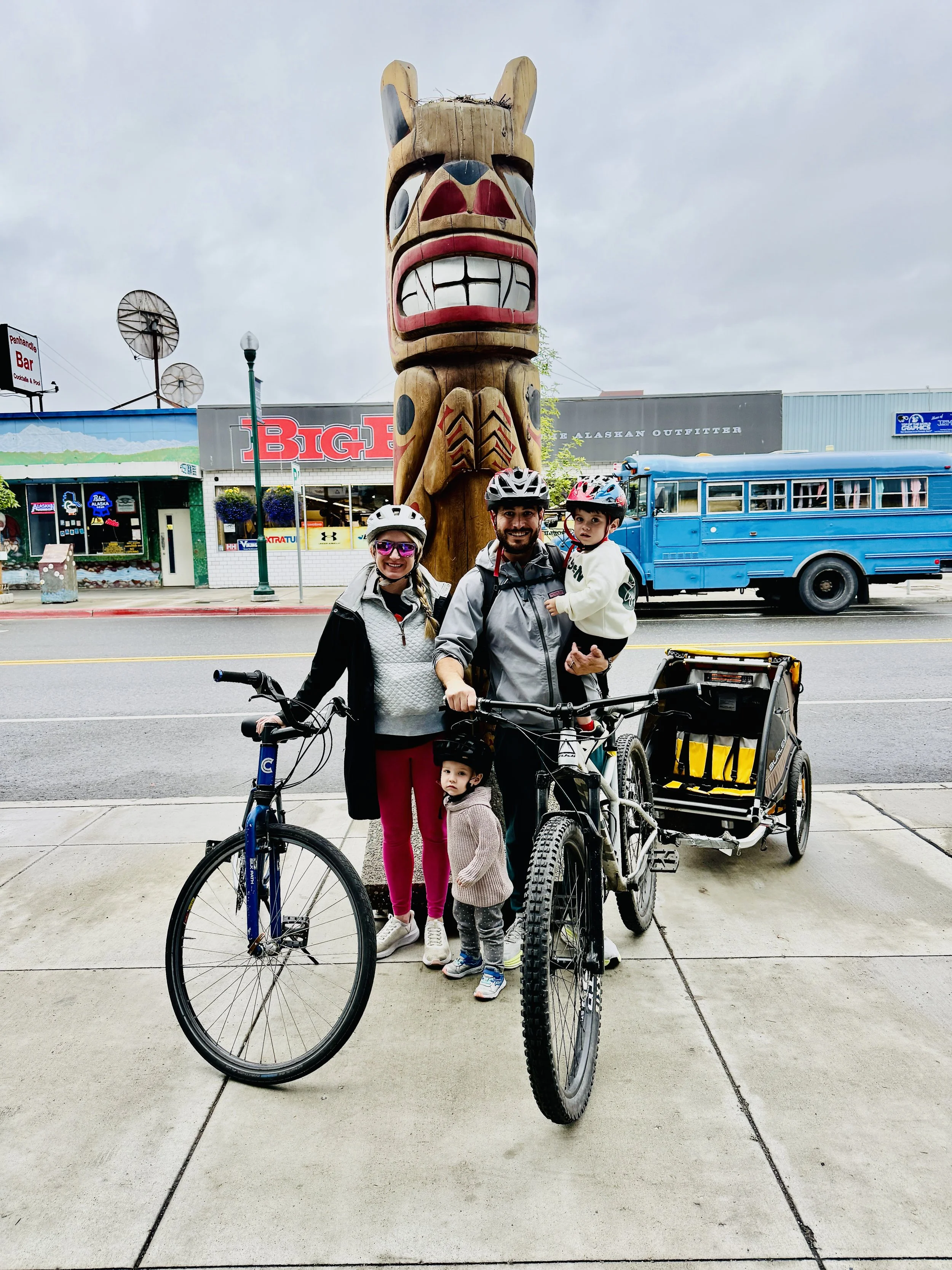 A family of four with two children, all wearing helmets, standing on a sidewalk in front of a large wooden totem pole with a totemic face carvings. The family is smiling, with one child sitting on a bicycle and the other child in their father's arms. Behind them are stores, a blue bus, and a cloudy sky.