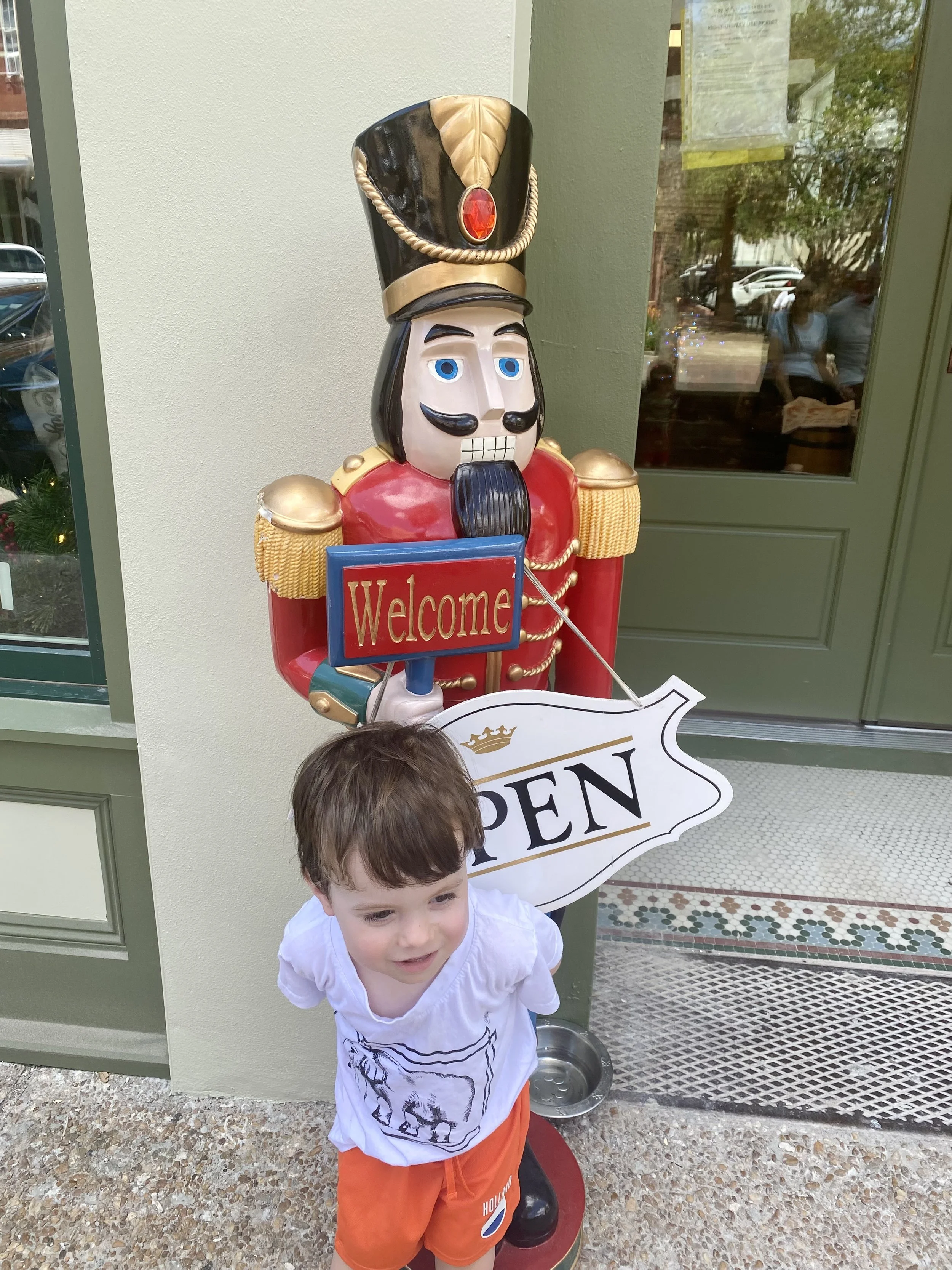 A young boy with brown hair and a white shirt is standing outside in front of a decorative nutcracker statue holding a red 'Welcome' sign. The nutcracker is dressed in a red and gold uniform with a black hat topped with a red gem. The boy appears to be smiling and is near a green door with a window, which reflects some trees and people inside a building.